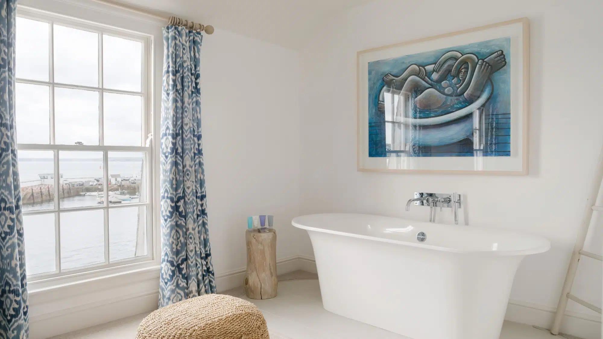 A free-standing white bathtub below a window looking out over a harbour full of moored boats, with patterned blue curtains and a framed painting on the wall.
