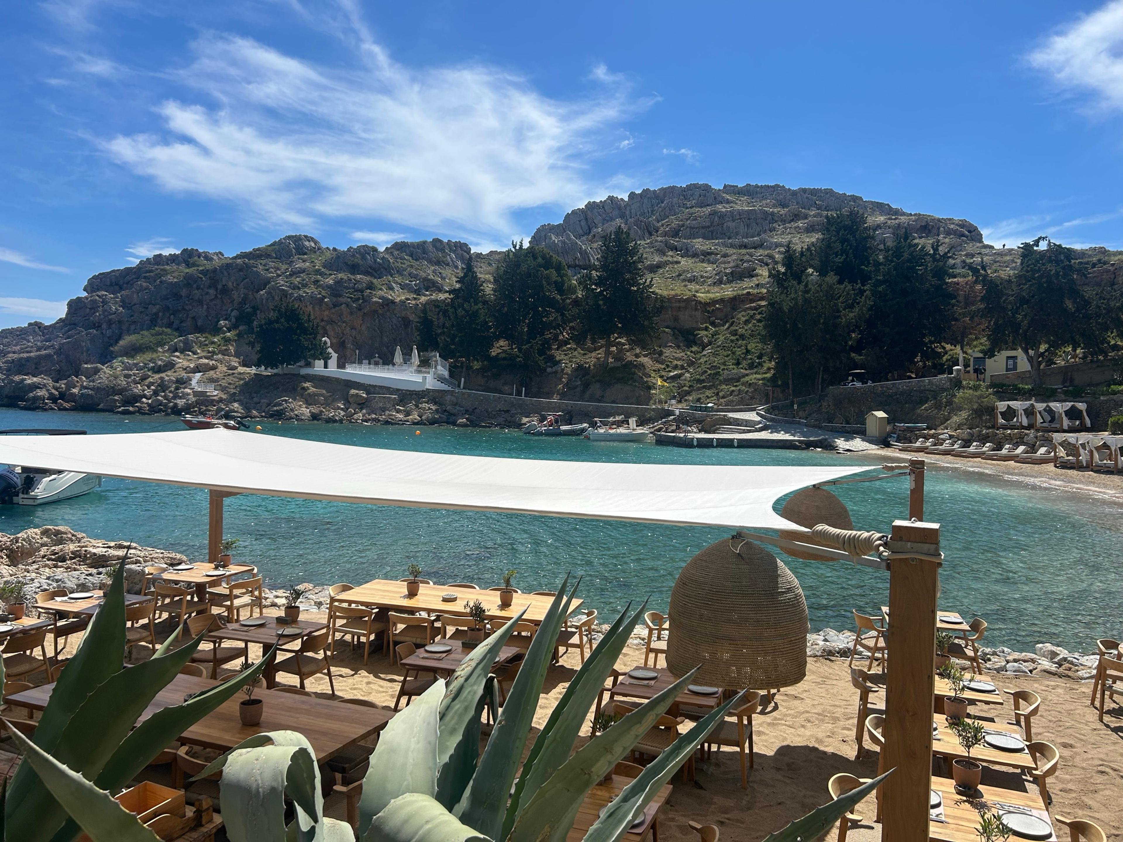 Tables laid on the sand under a white shade sail at St Paul's Bay, Lindos, with turquoise water, a small white chapel and rocky hills behind