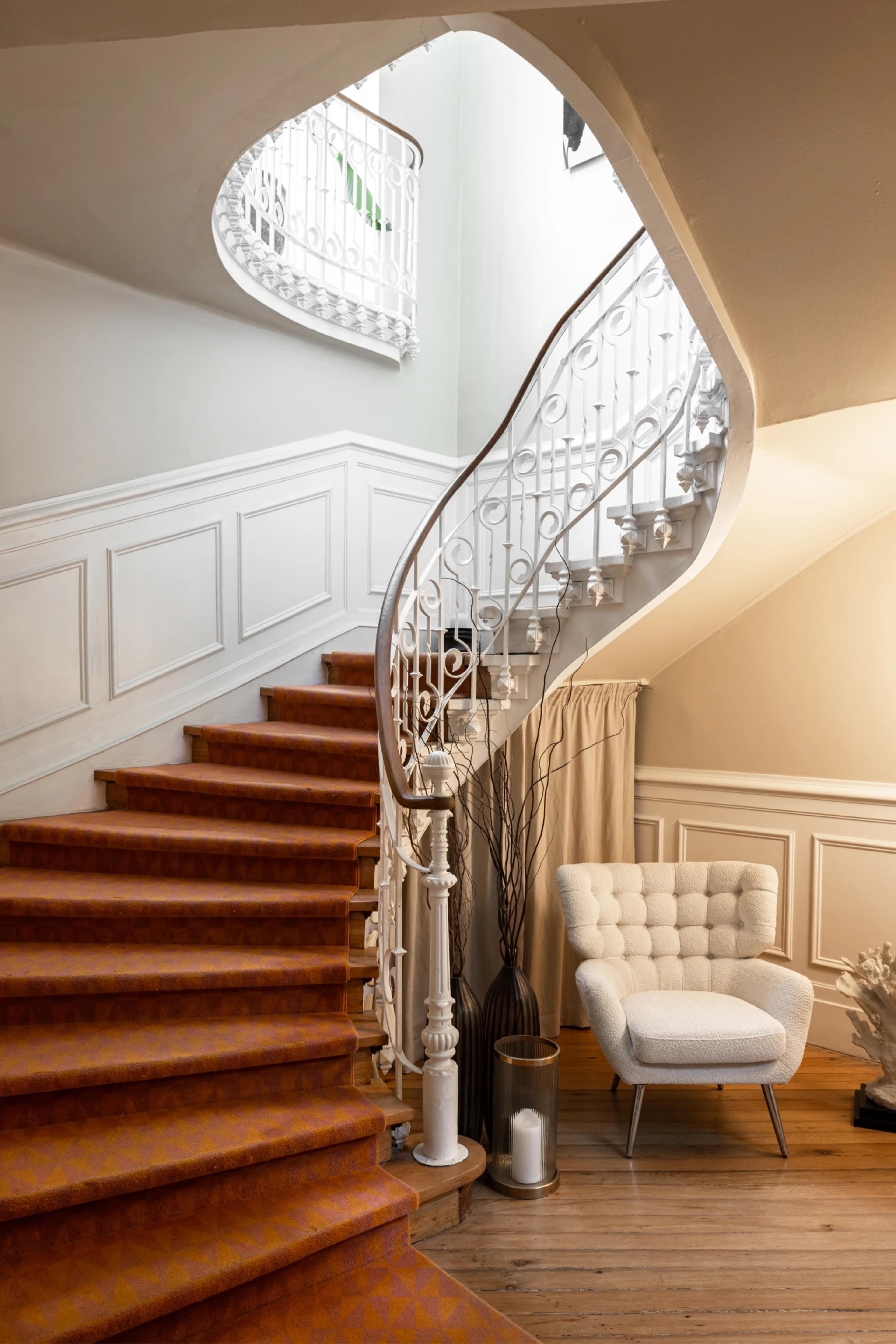A curved staircase with a white wrought-iron balustrade and a timber handrail rises beside a wall of white panelling, with orange-brown carpet on the steps and a white armchair at the foot.