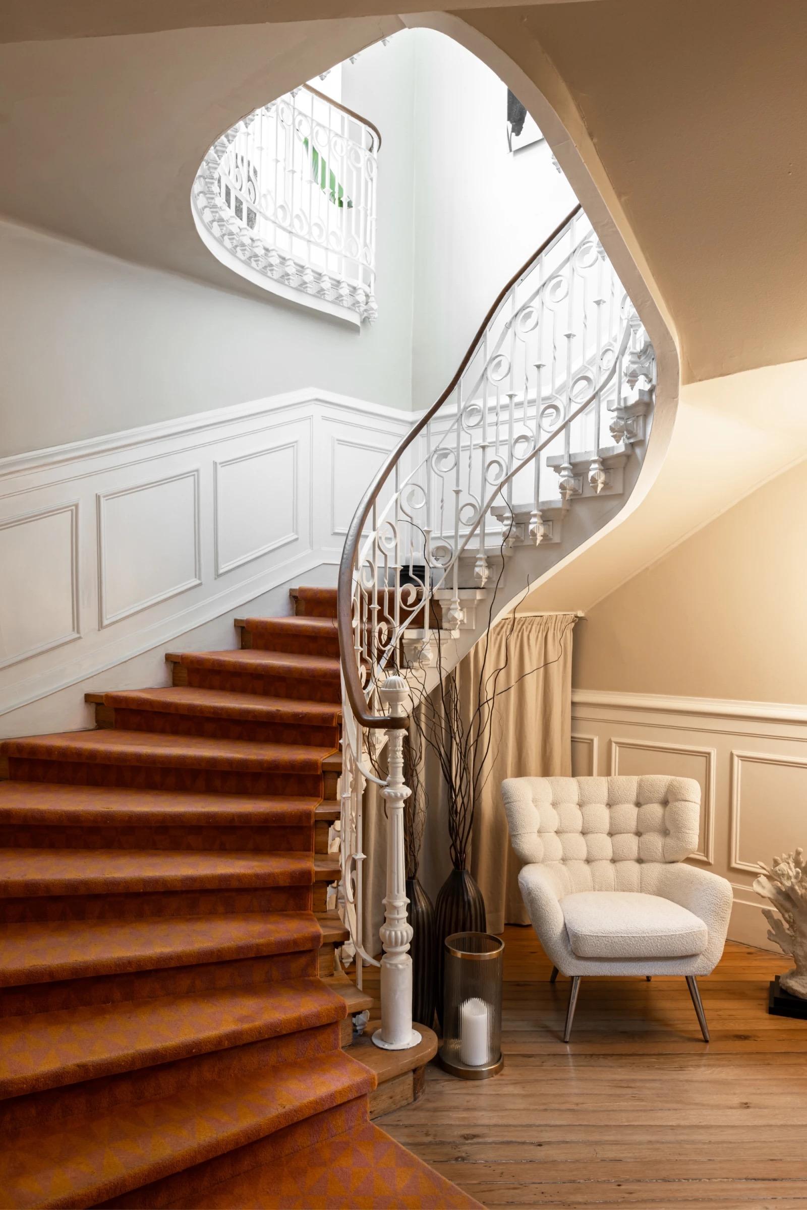 A curved staircase with a white wrought-iron balustrade and a timber handrail rises beside a wall of white panelling, with orange-brown carpet on the steps and a white armchair at the foot.