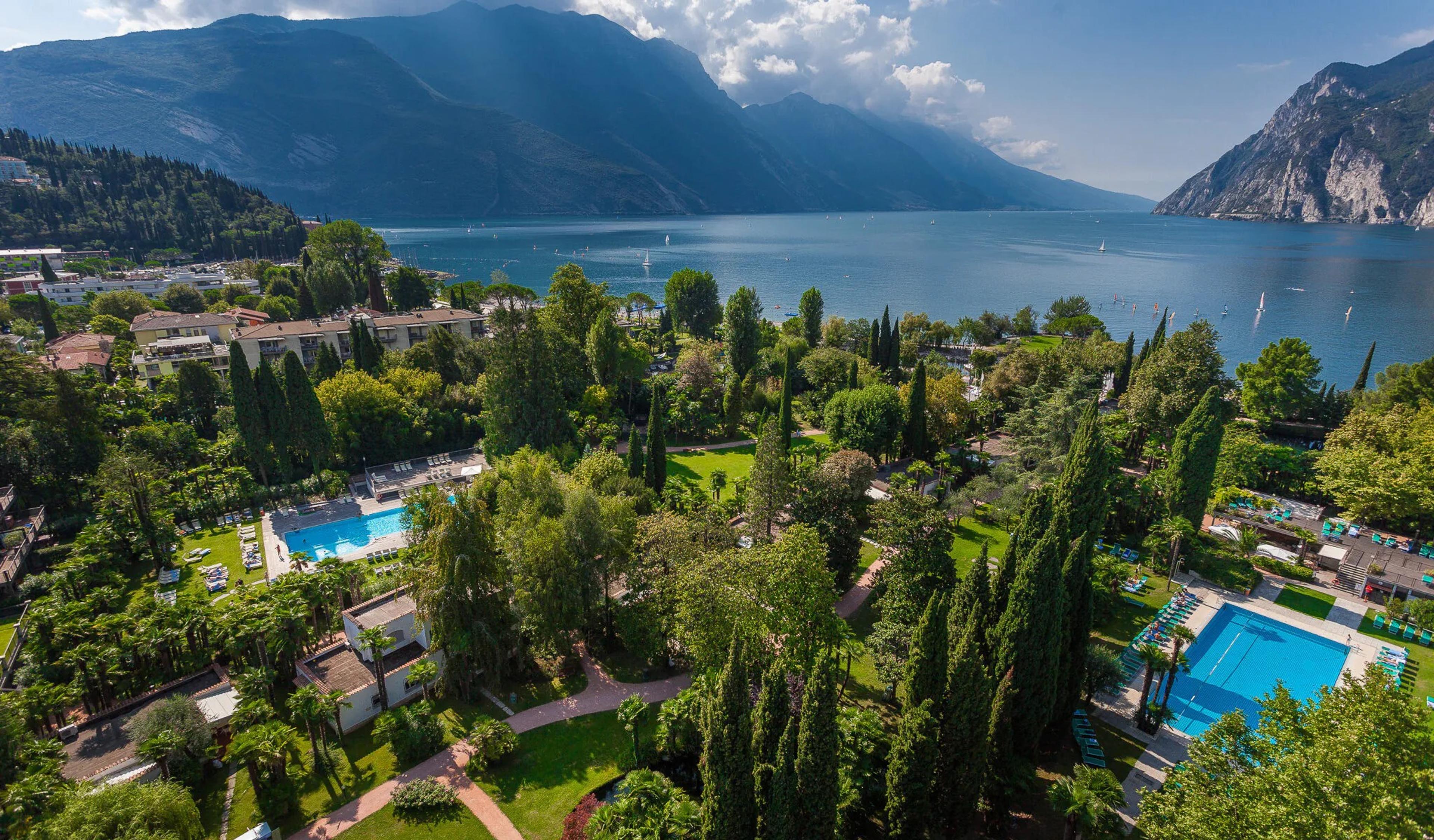 Aerial view of a hotel garden on the shore of Lake Garda, with two swimming pools among lawns and trees, white buildings at the left, and mountains rising over the water.