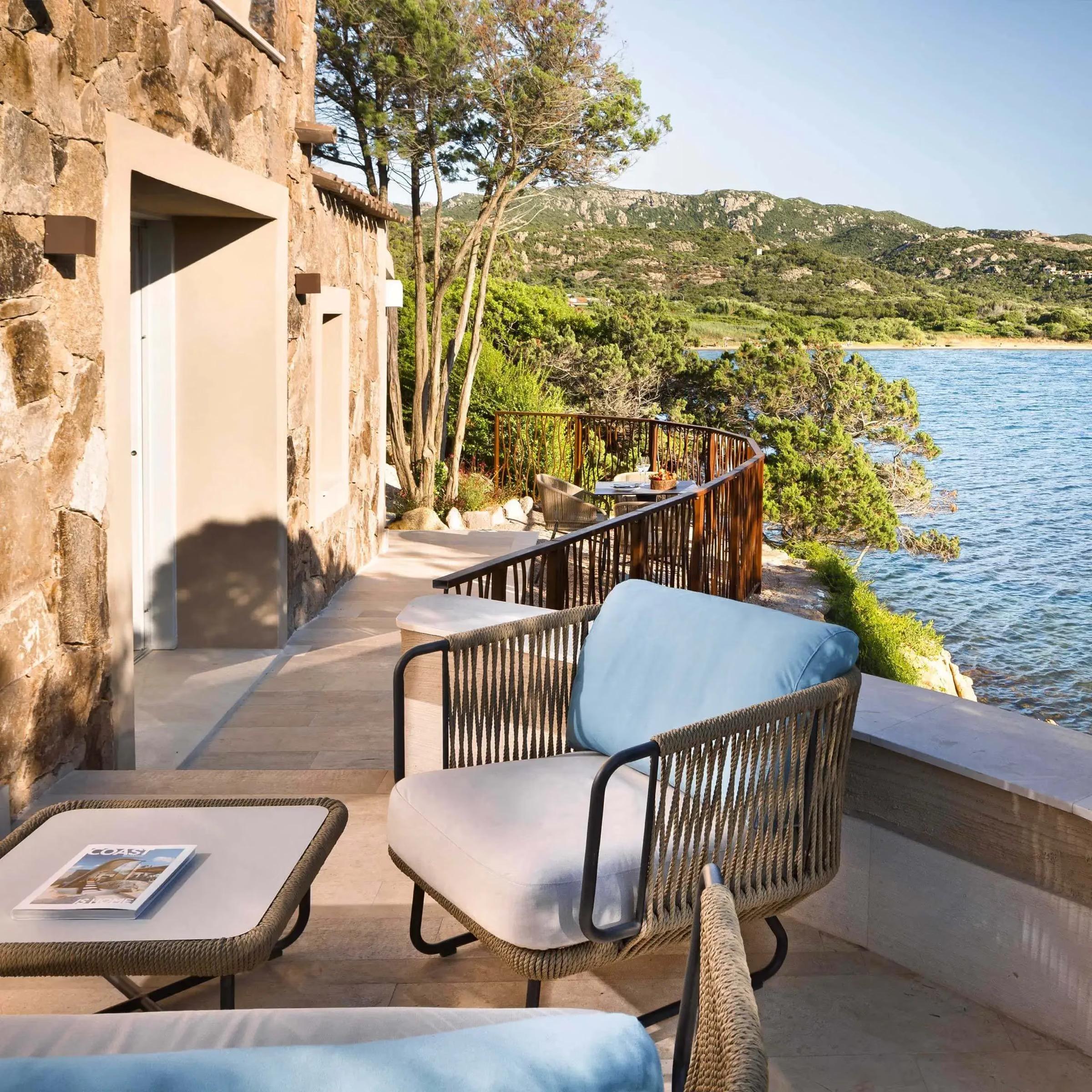 A terrace with woven outdoor chairs and a side table looks over coastal pine and rock to the open sea and hills beyond.
