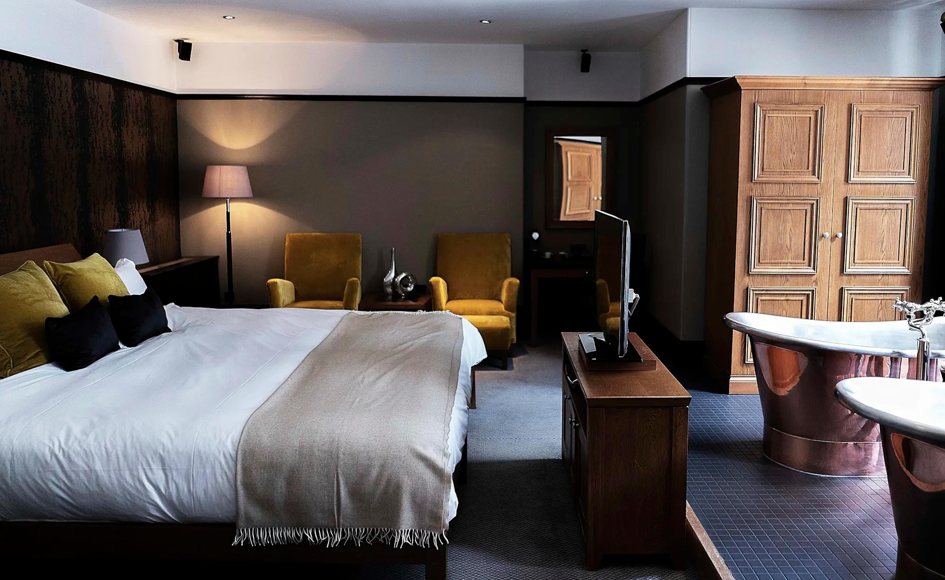 A bedroom with a king bed on the left, two yellow armchairs and a flat-screen television, and two copper bathtubs on a tiled platform beside tall windows on the right.