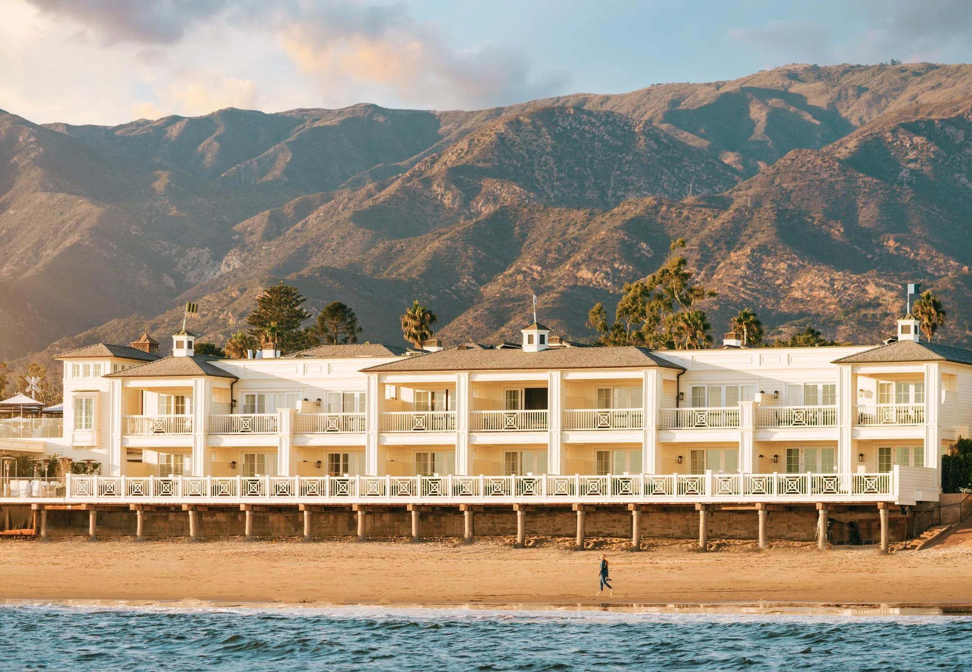A three storey white clapboard hotel built on pilings over a sandy beach, with mountains behind it and a person walking along the shore.