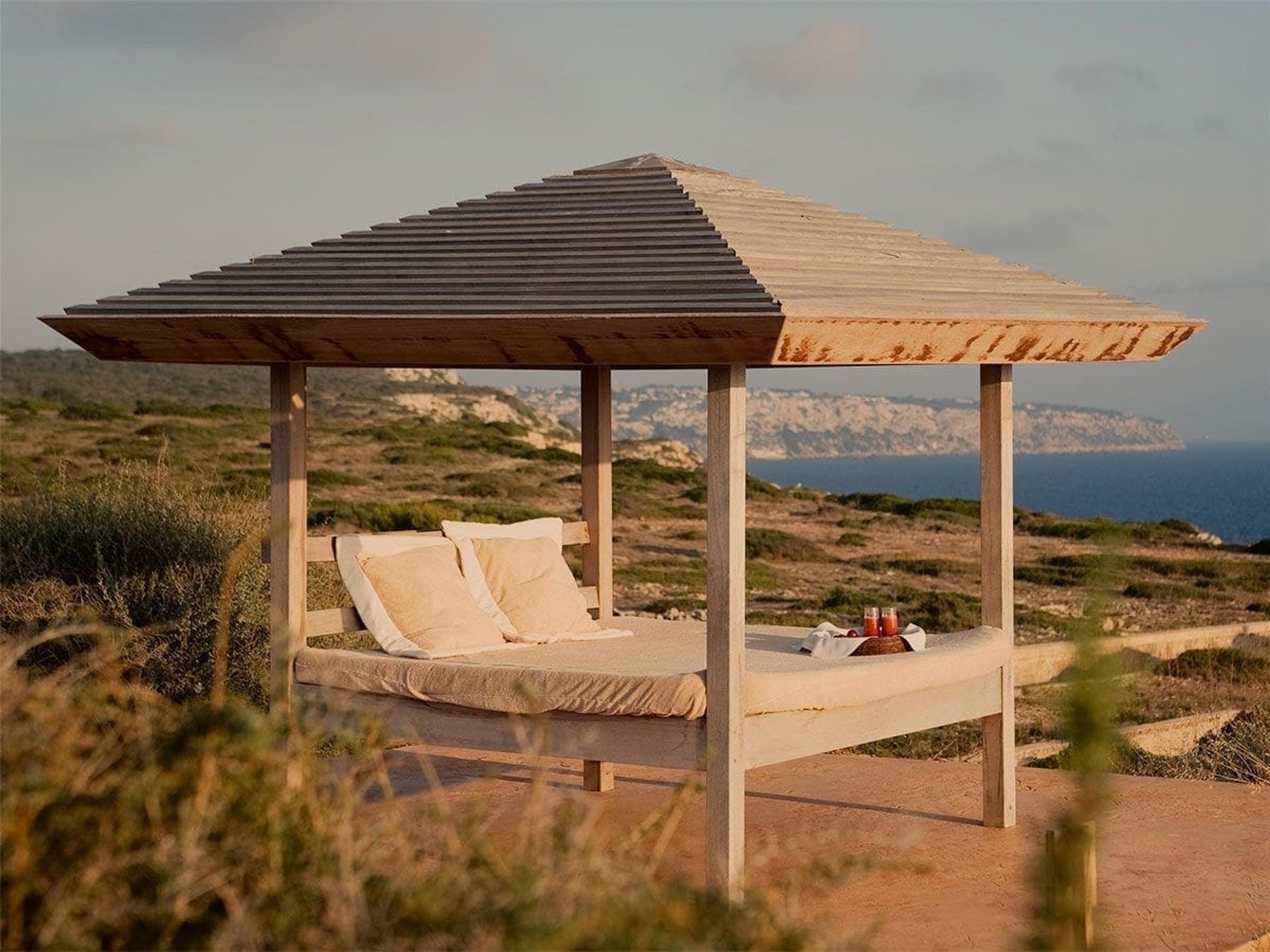 A wooden daybed under a pitched wooden canopy on a clifftop, with cushions and a tray of drinks, overlooking the coastline and the sea.