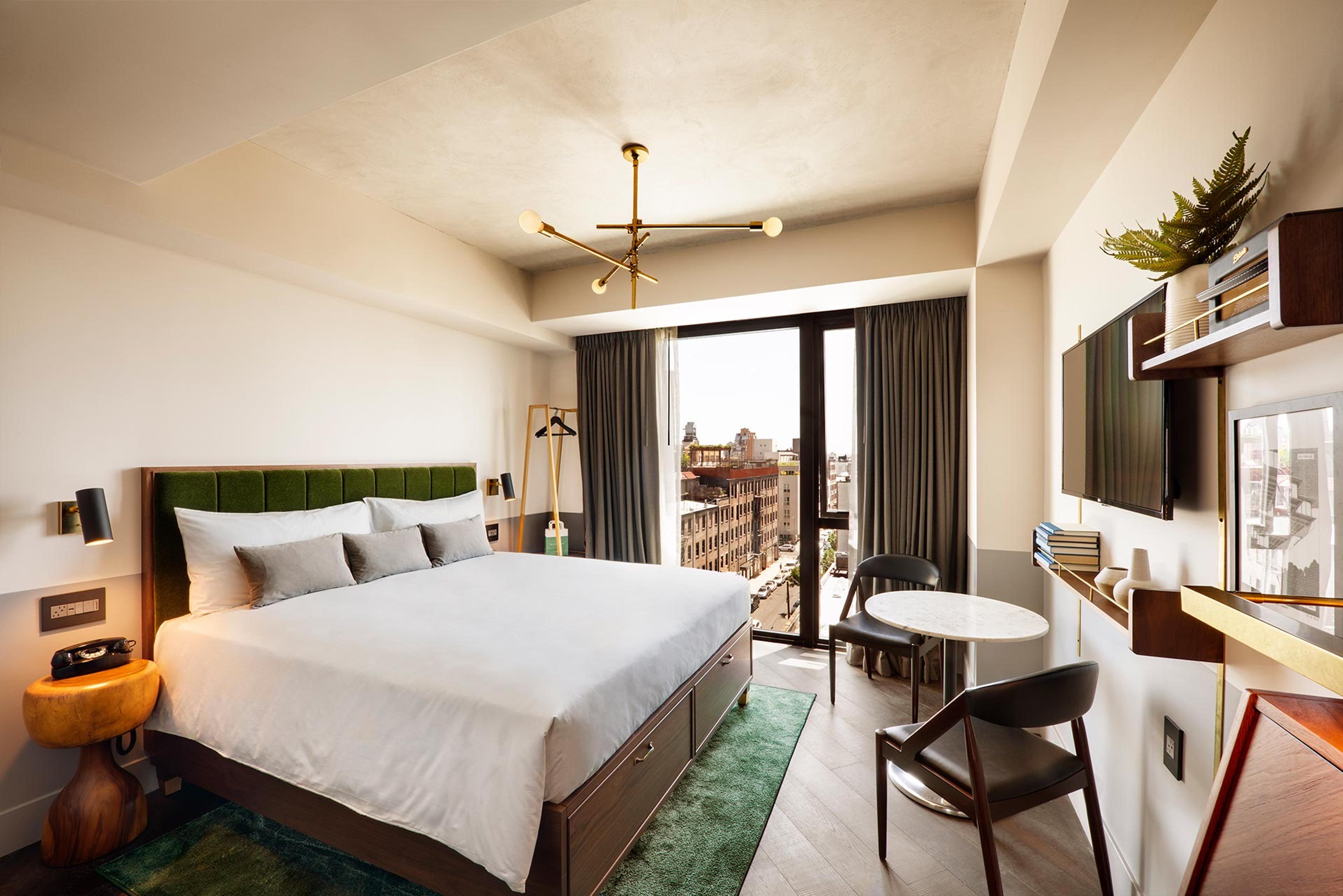a king bed, a round table with two chairs, and an open balcony door looking onto a street, with a wall-mounted TV and a brass branched light fitting