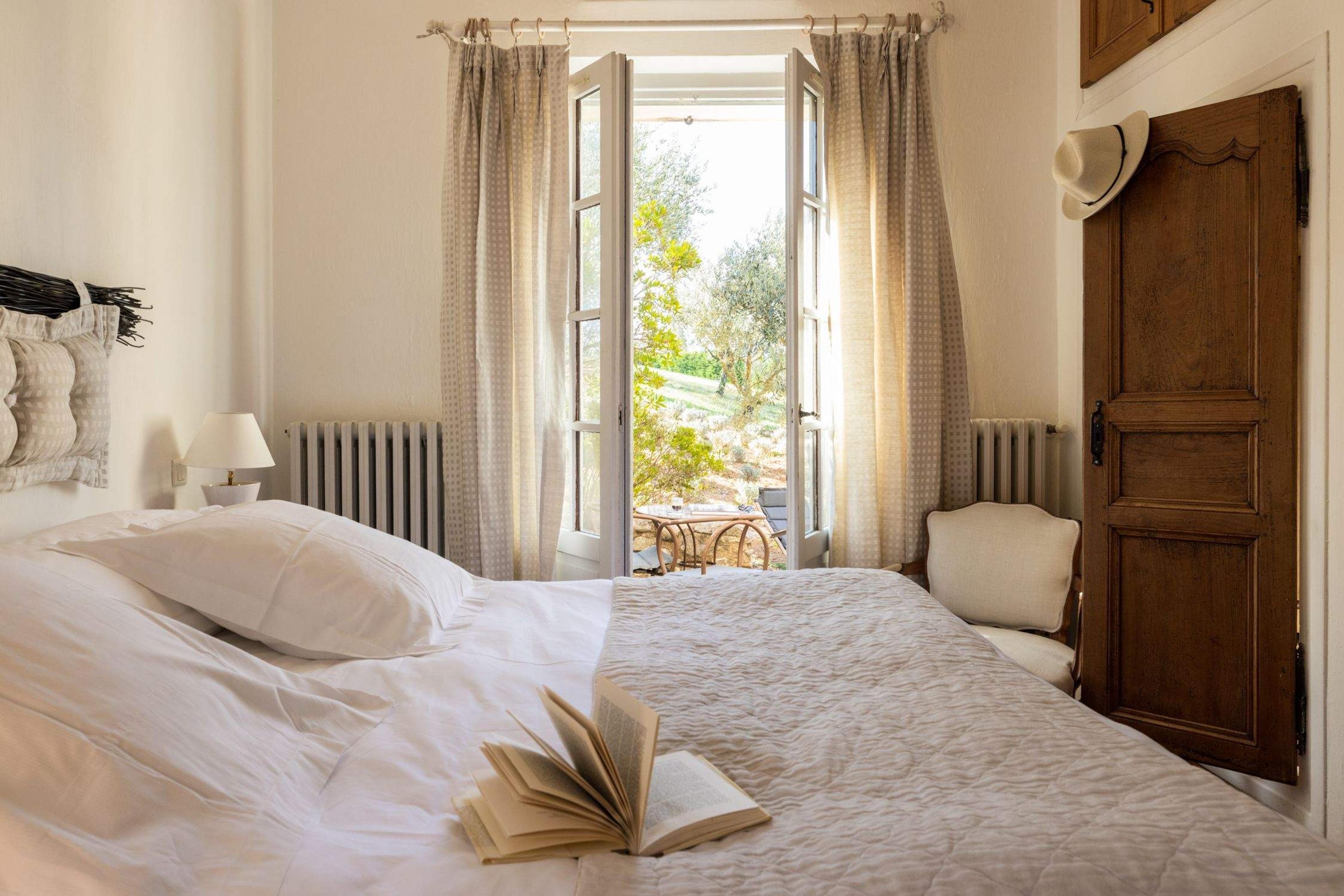 A bedroom with a white-linened bed, an open book, French doors open to olive trees beyond, and a straw hat hanging on the door.