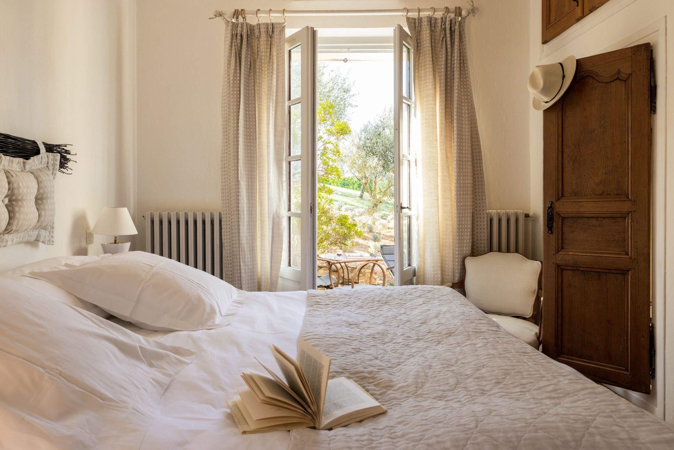 A bedroom with a white-linened bed, an open book, French doors open to olive trees beyond, and a straw hat hanging on the door.