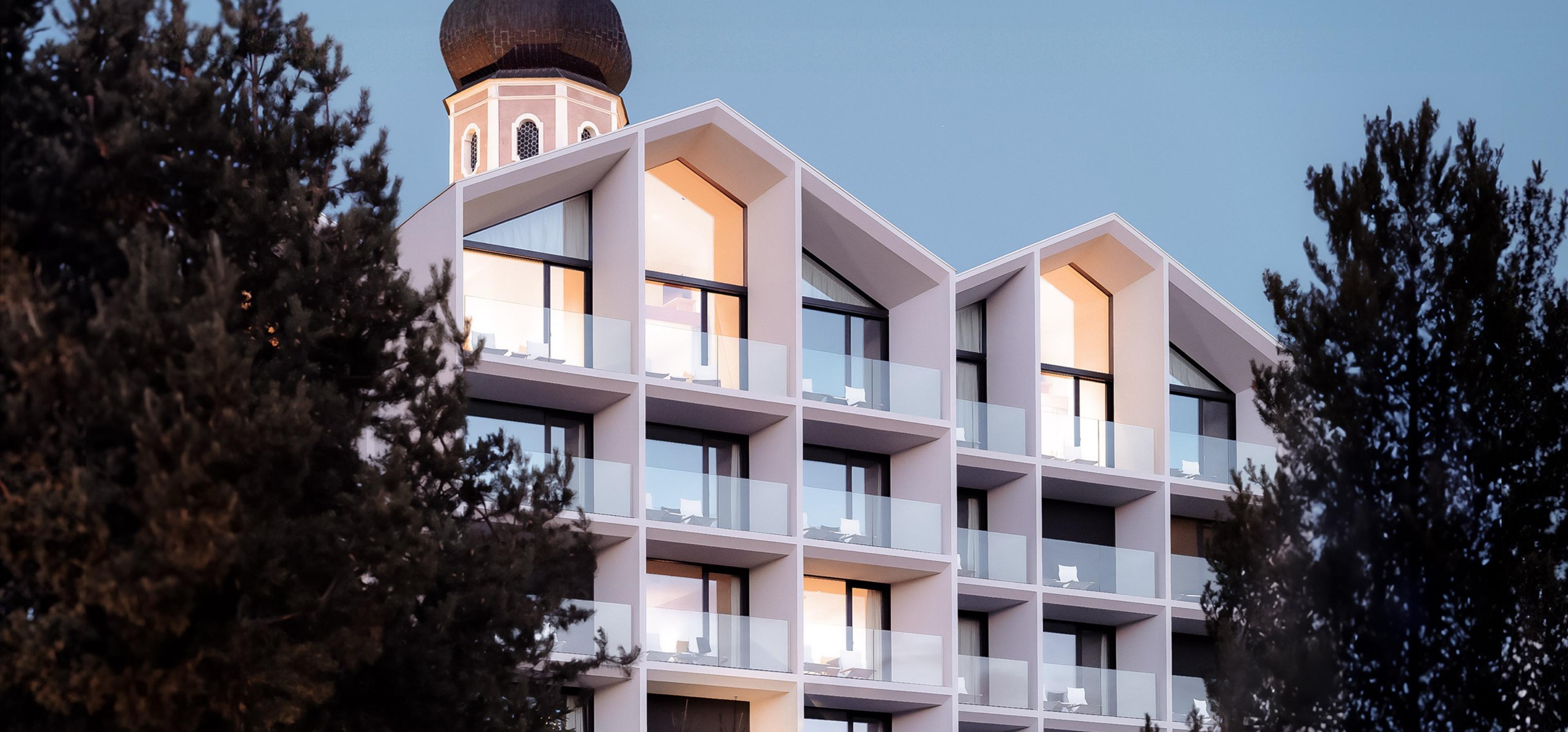 The hotel's pitched, gabled facade at dusk, lit from inside and framed by pine trees, with a village church tower behind it.