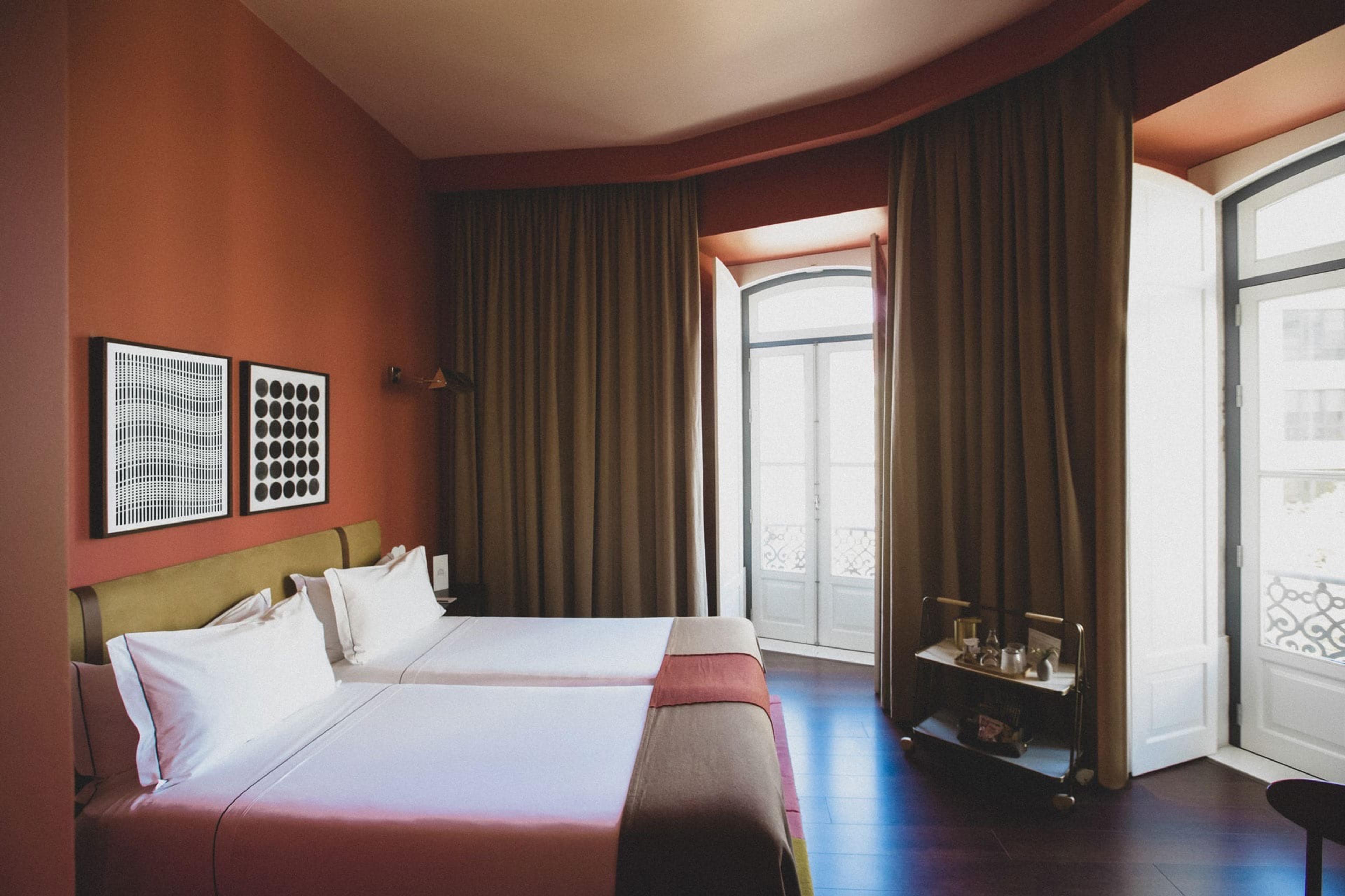 A bedroom with terracotta walls, two made-up beds, brown curtains and tall windows opening onto a balcony.