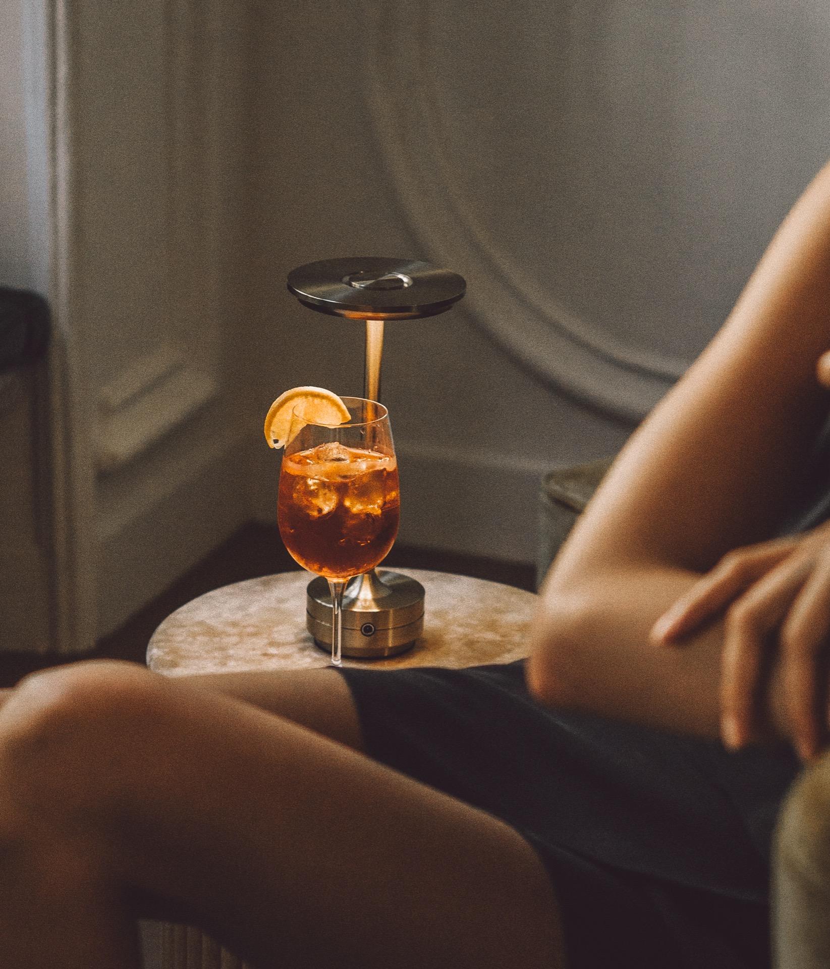 An Aperol spritz with an orange wheel sits on a small marble side table beside a seated person's bare legs, lit by a warm lamp.