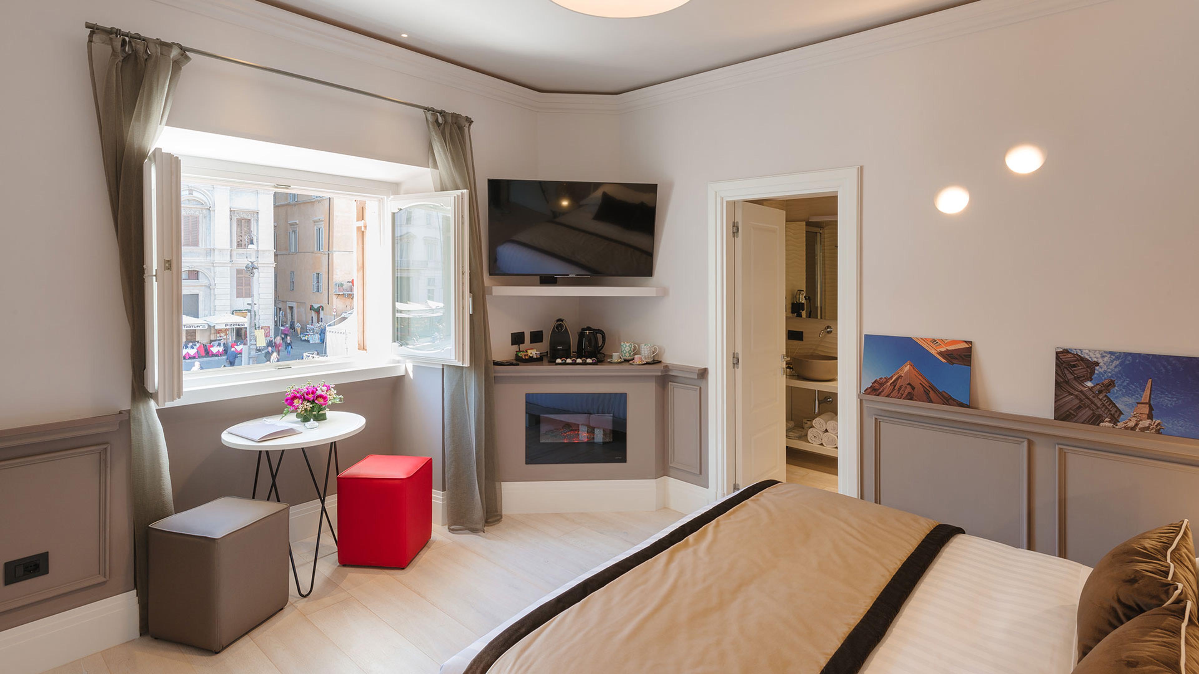 A corner bedroom with an open window looking onto a busy sunlit street, a small round table with flowers, a wall-mounted television, and an electric fireplace.
