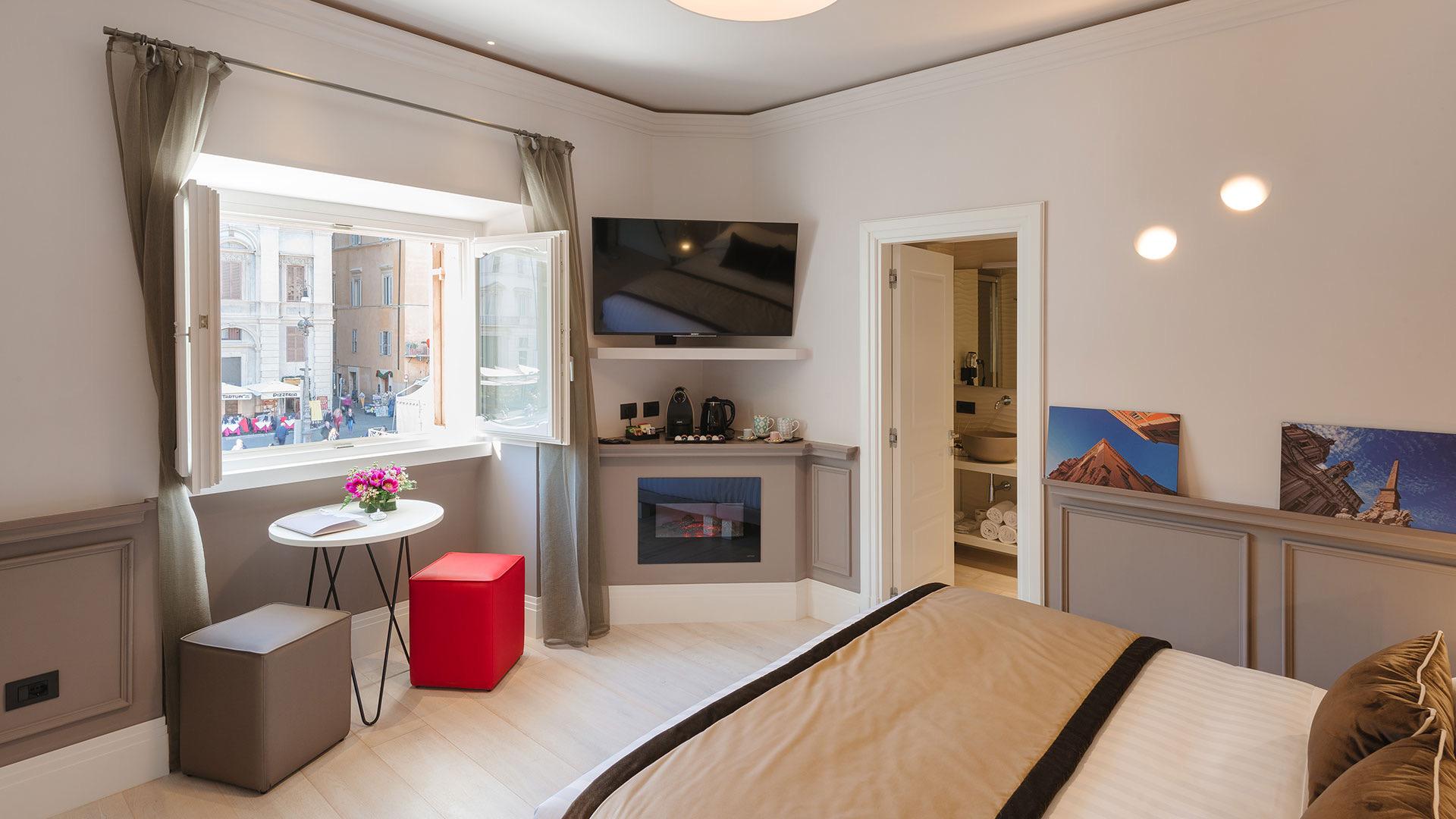 A corner bedroom with an open window looking onto a busy sunlit street, a small round table with flowers, a wall-mounted television, and an electric fireplace.