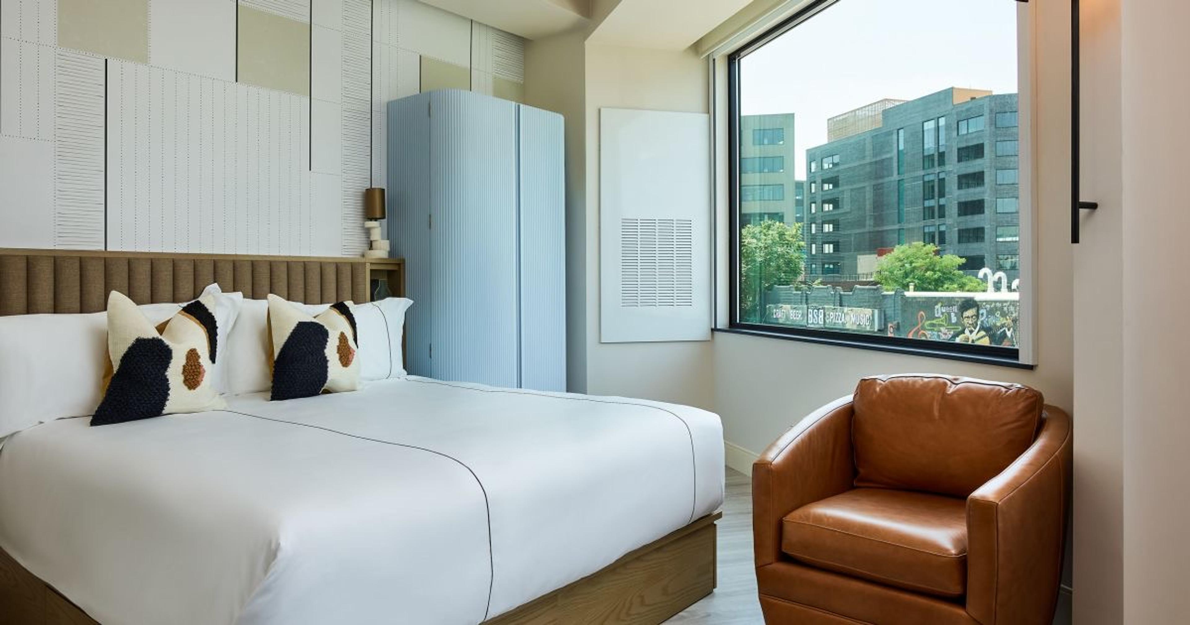 A hotel bedroom with a brown leather armchair, a white bed with patterned pillows, and a large window looking onto a building with street-level graffiti.
