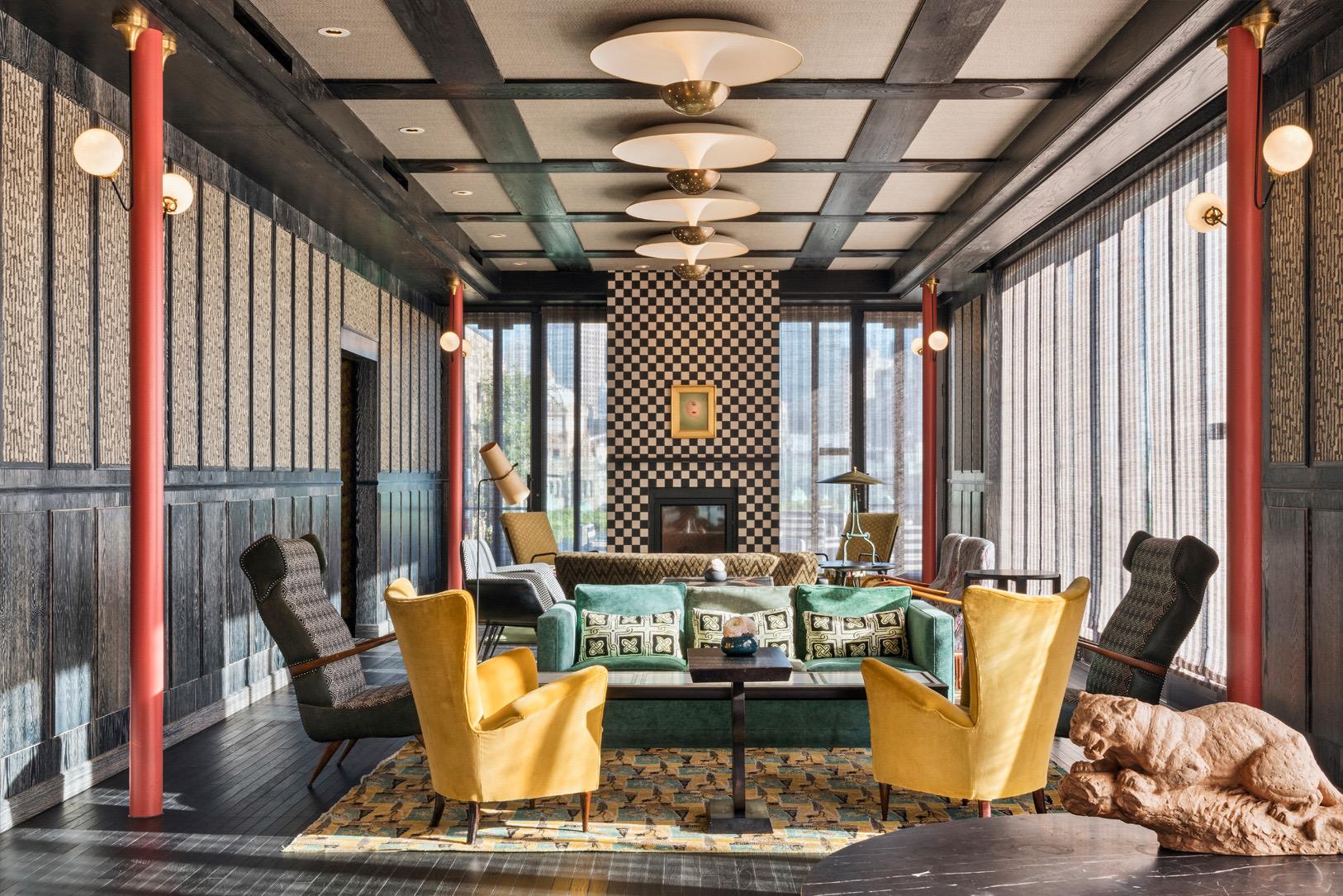 A lounge with mustard-yellow wingback chairs, a green sofa, a checkerboard-tiled fireplace wall and a stone sculpture of a reclining animal in the foreground.