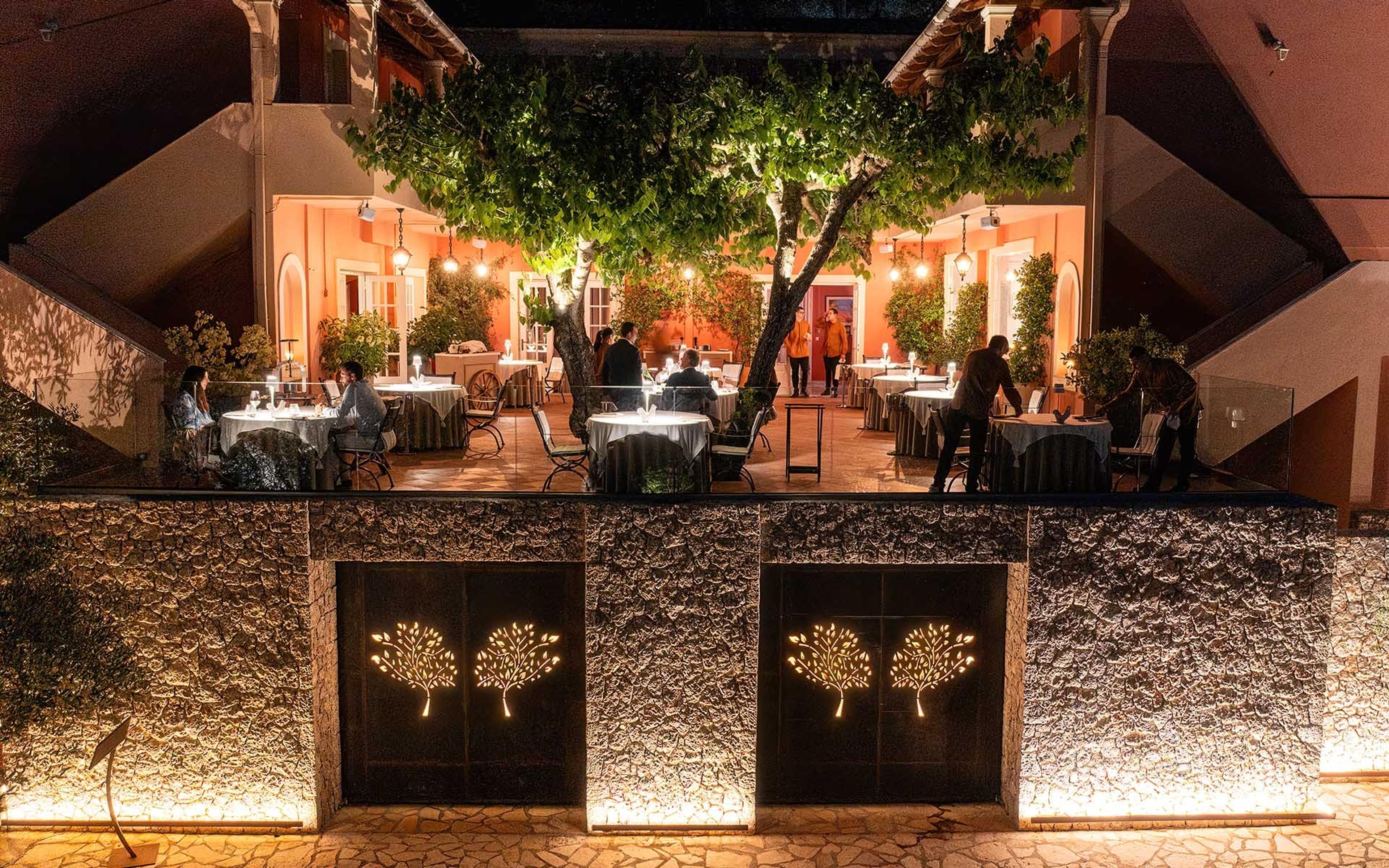 Tables lit at night in a courtyard under a spreading tree, behind a stone wall