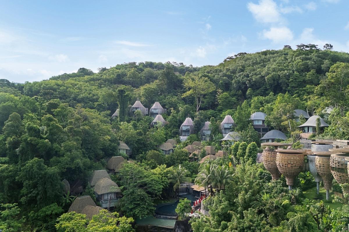 An aerial view of the resort's cone-roofed and basket-shaped villas spread across a forested hillside under a blue sky.