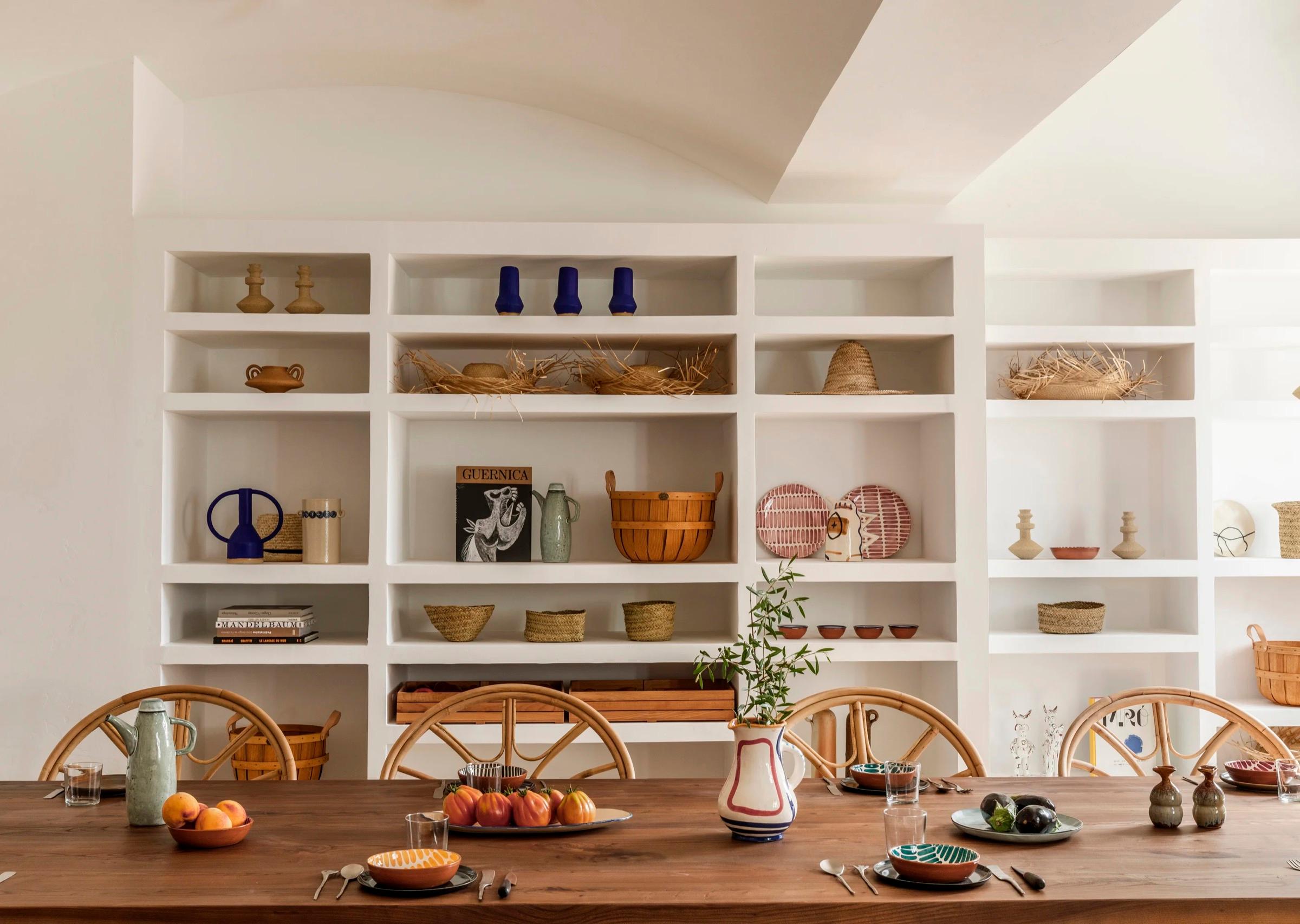 A wooden table set with bowls of fruit and ceramics in front of white built-in shelving holding baskets, pottery, and books.
