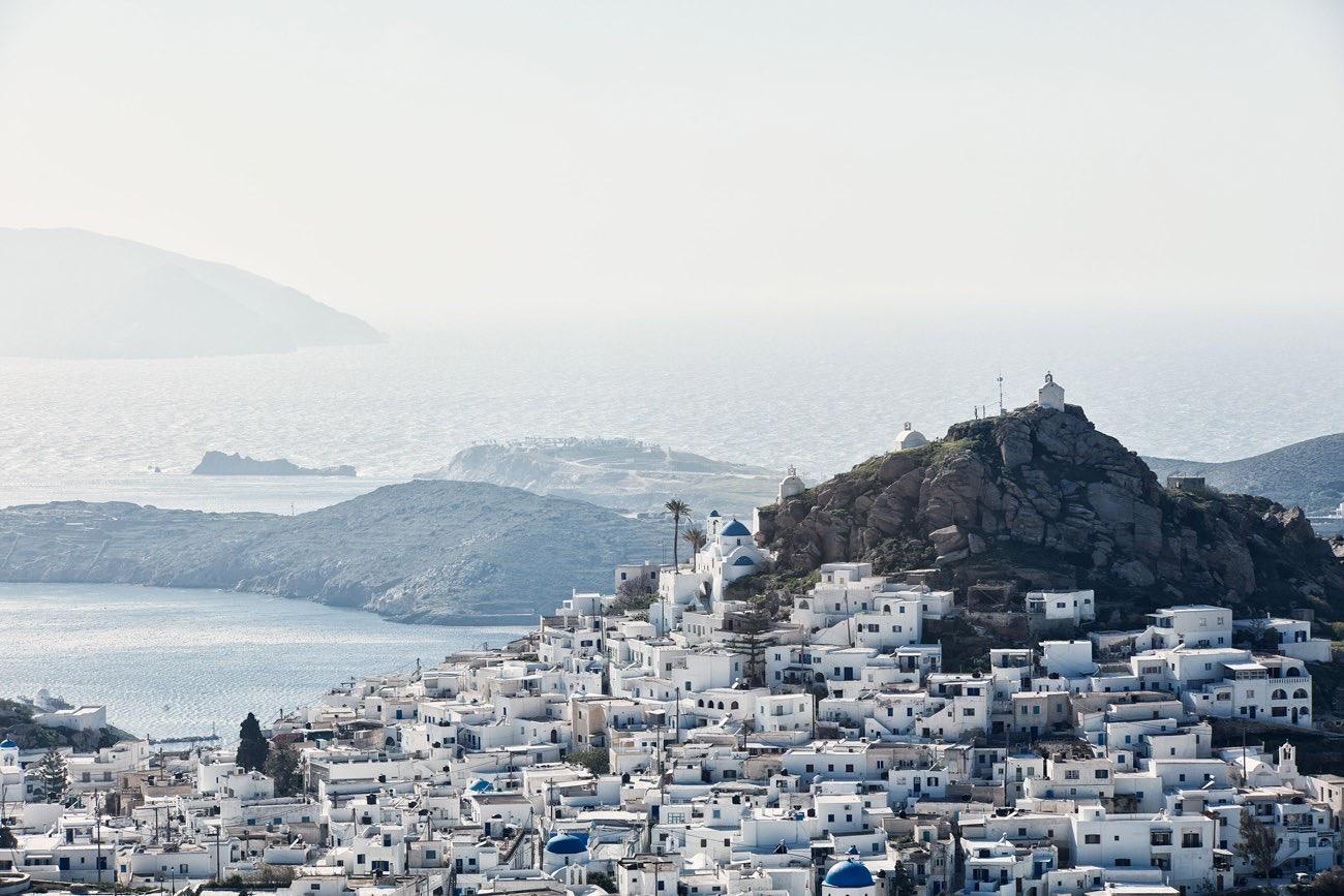 A white hillside village with blue-domed churches and a rocky headland, with the sea and islands hazy below.