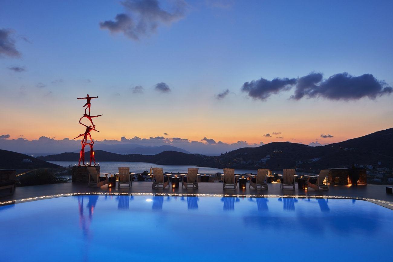 A pool lit blue at dusk, with loungers and lanterns along its edge, a red sculpture of acrobats on a plinth, and hills and sea beyond.