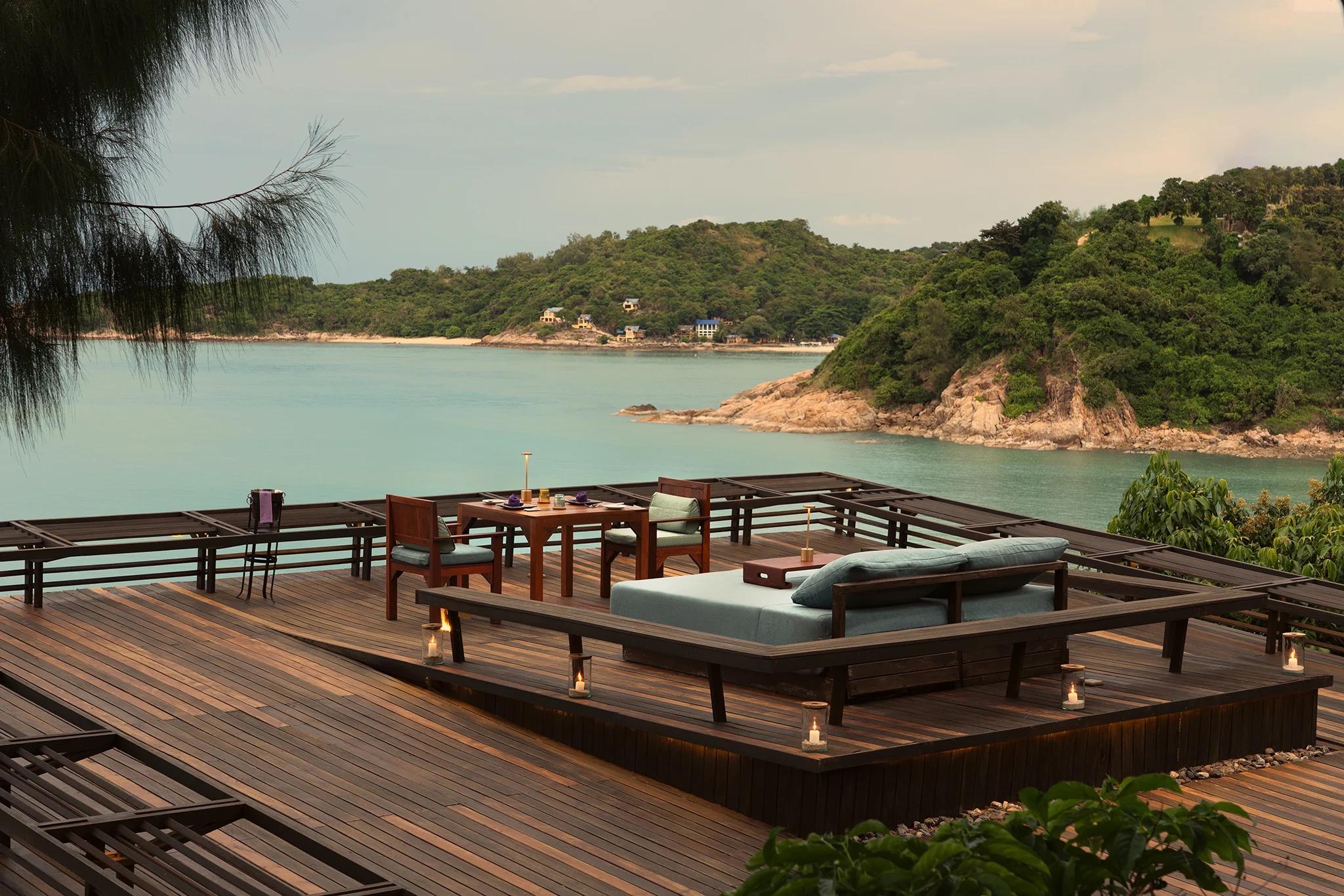 A wooden deck with a dining table, chairs and a daybed looks out over a calm bay toward tree-covered hills and a scatter of houses on the far shore.