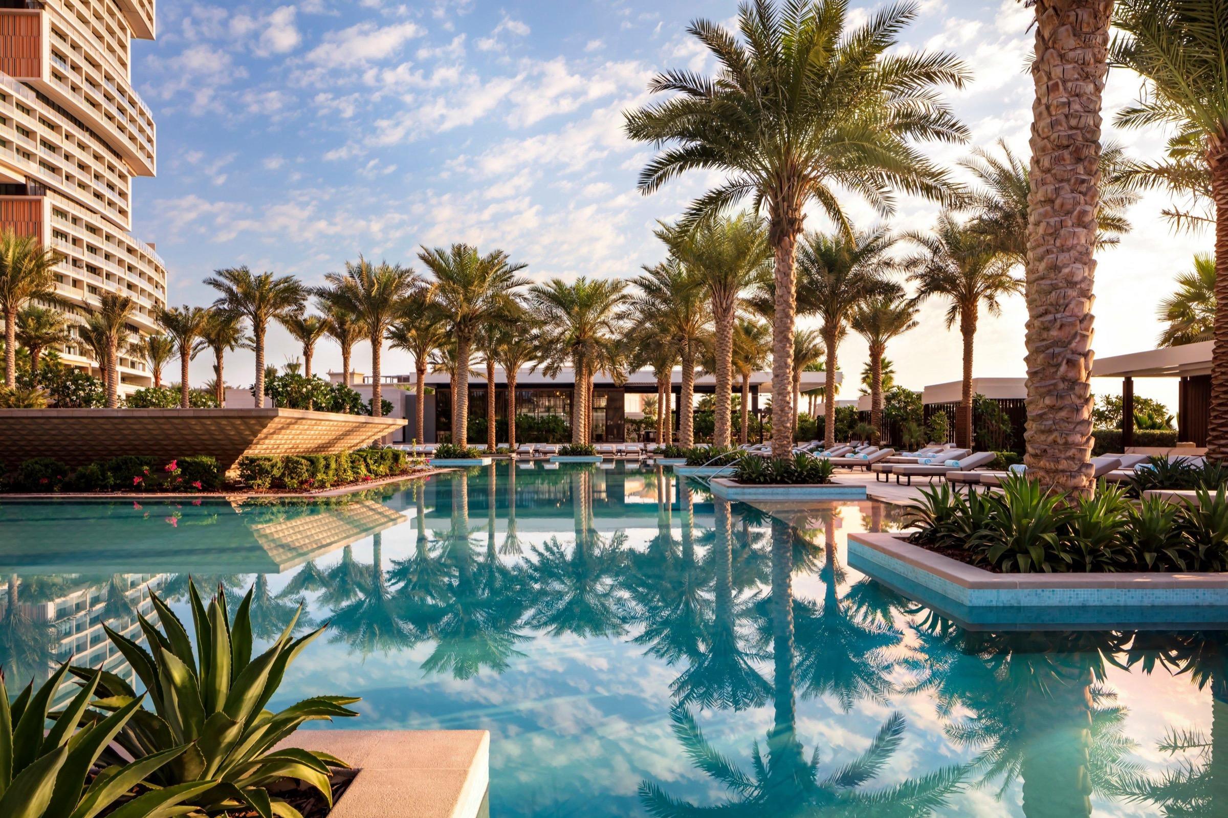 A long infinity pool lined with palm trees and sun loungers, with the hotel's tiered tower rising behind it.