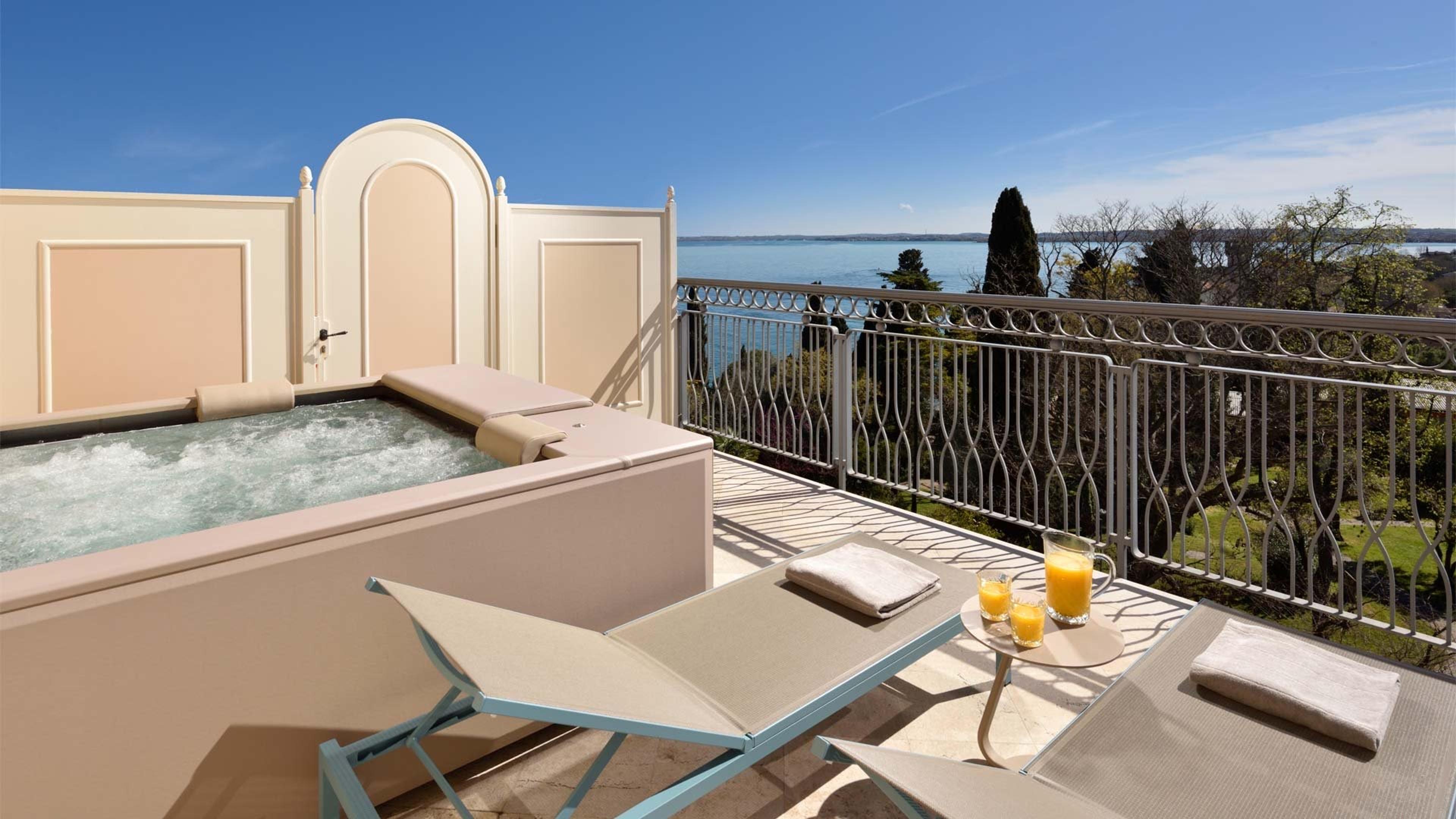 A private terrace with a built-in hot tub, two loungers and a jug of orange juice, overlooking a lake.
