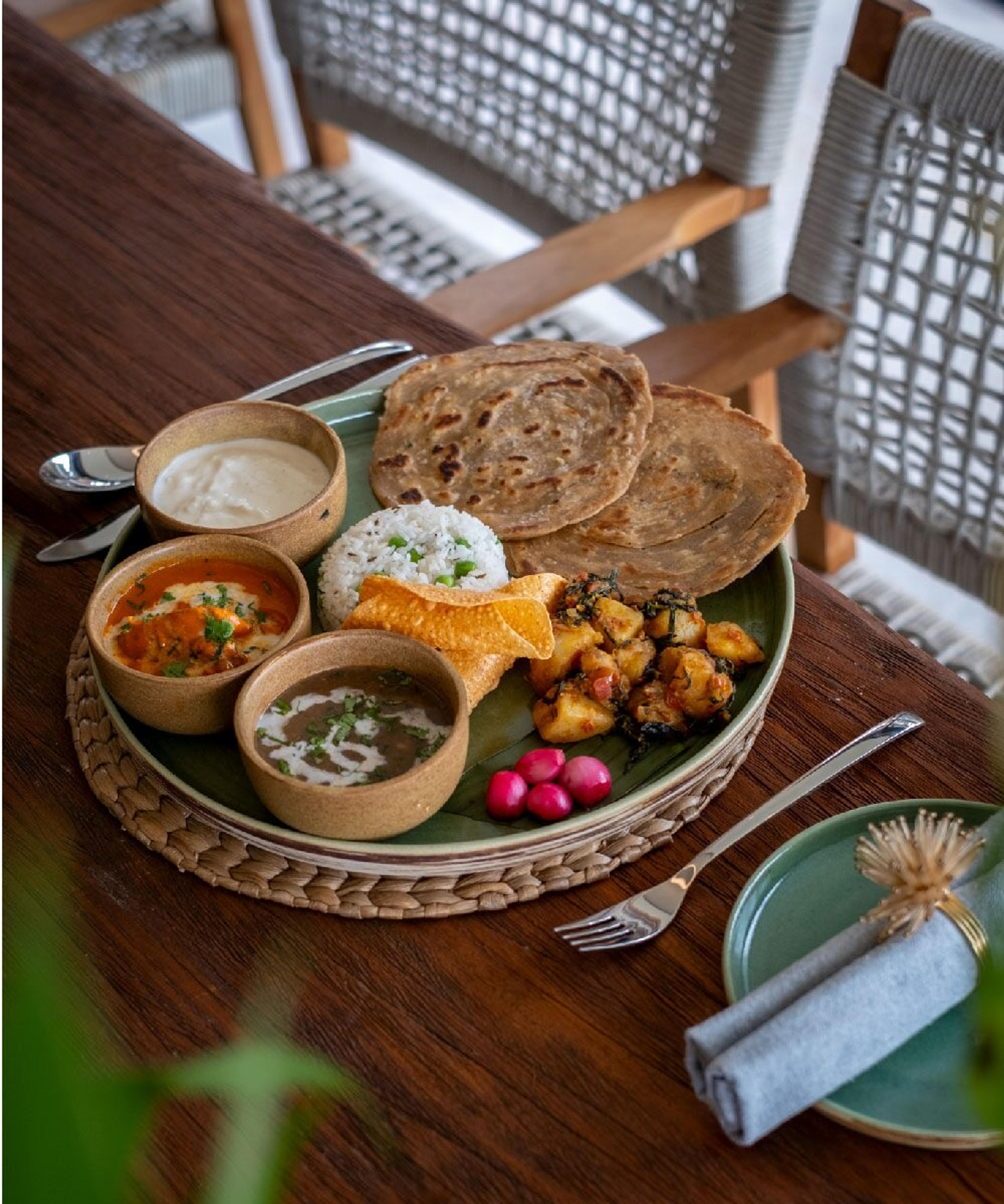A round thali tray holding rice, two flatbreads, four small bowls of curry and chutney, and a pappadum, on a wooden table.