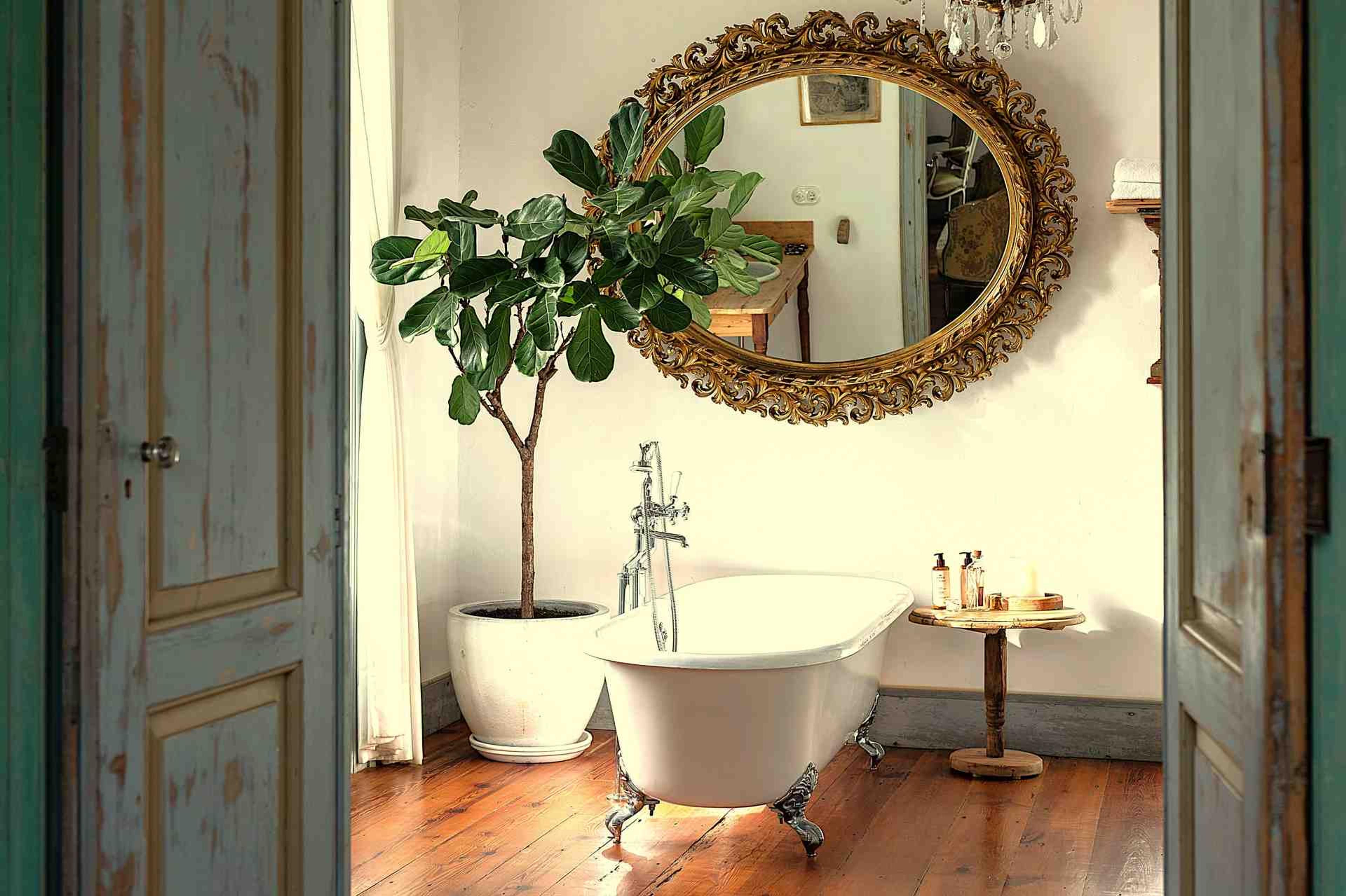 Seen through a pair of open doors, a freestanding bathtub beneath a large gilt oval mirror, with a leafy potted plant and a side table of toiletries.
