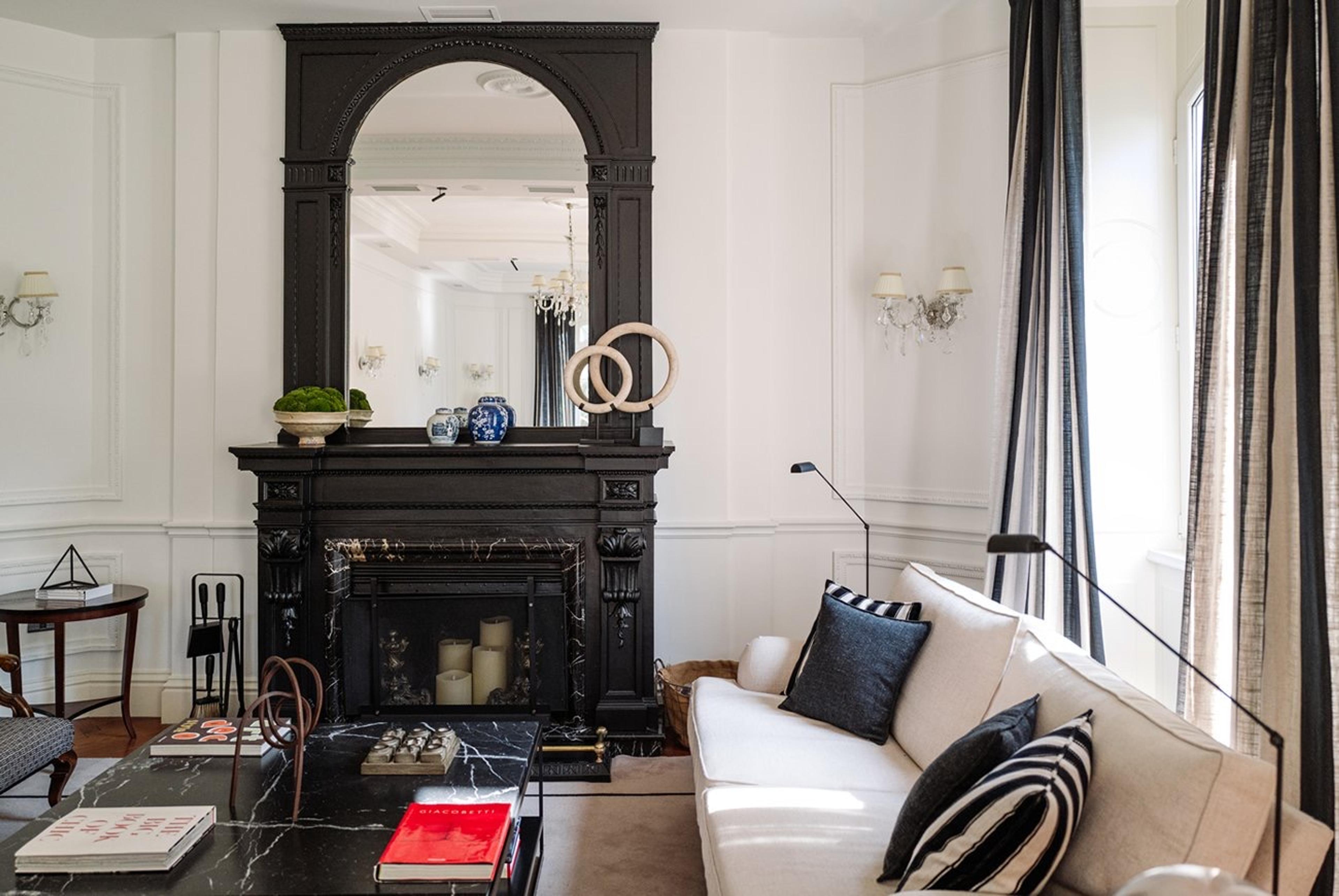 A sitting room with a tall black fireplace and mirror, a white sofa with striped and checked cushions, and floor lamps either side.