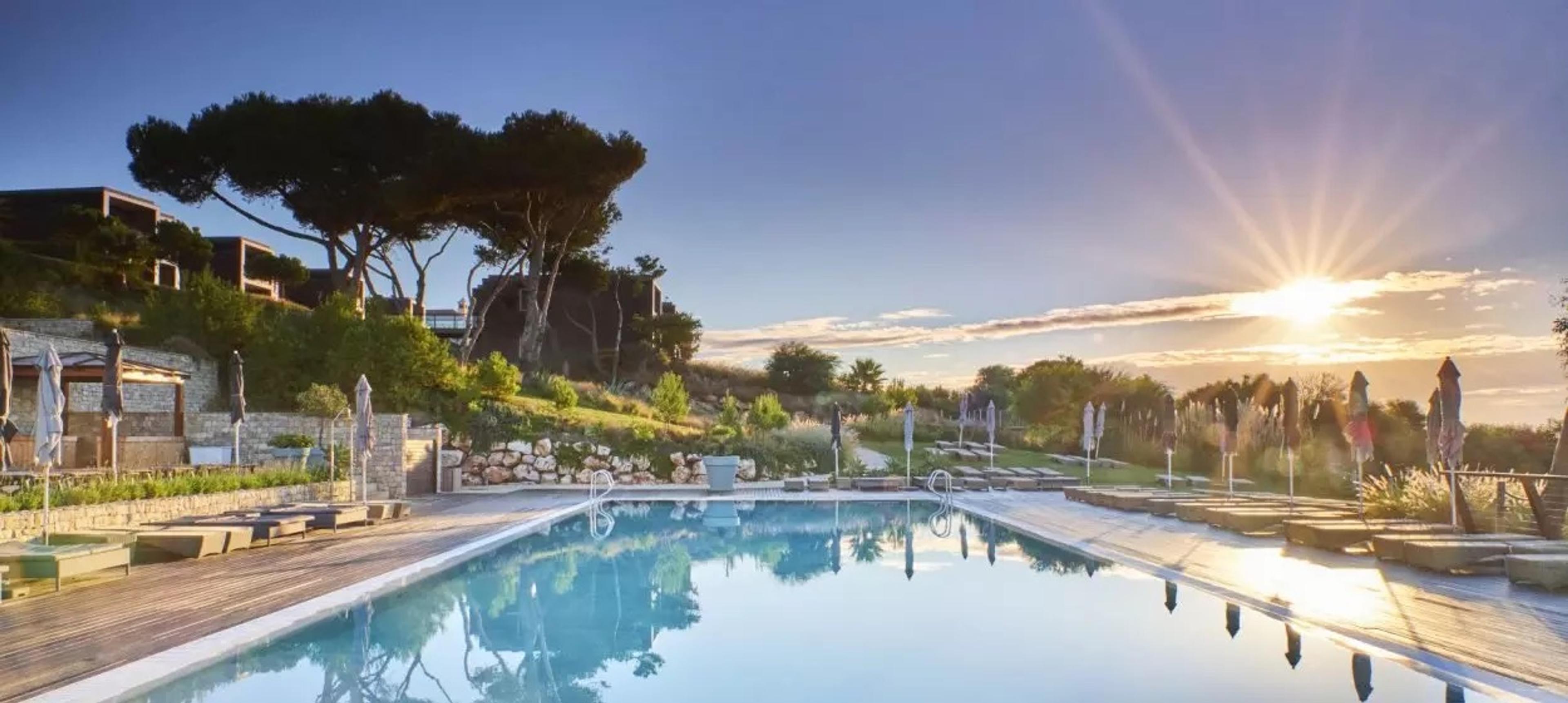 A pool at sunset with a stone wall, pine trees and loungers around it, and the low sun flaring over the hills.