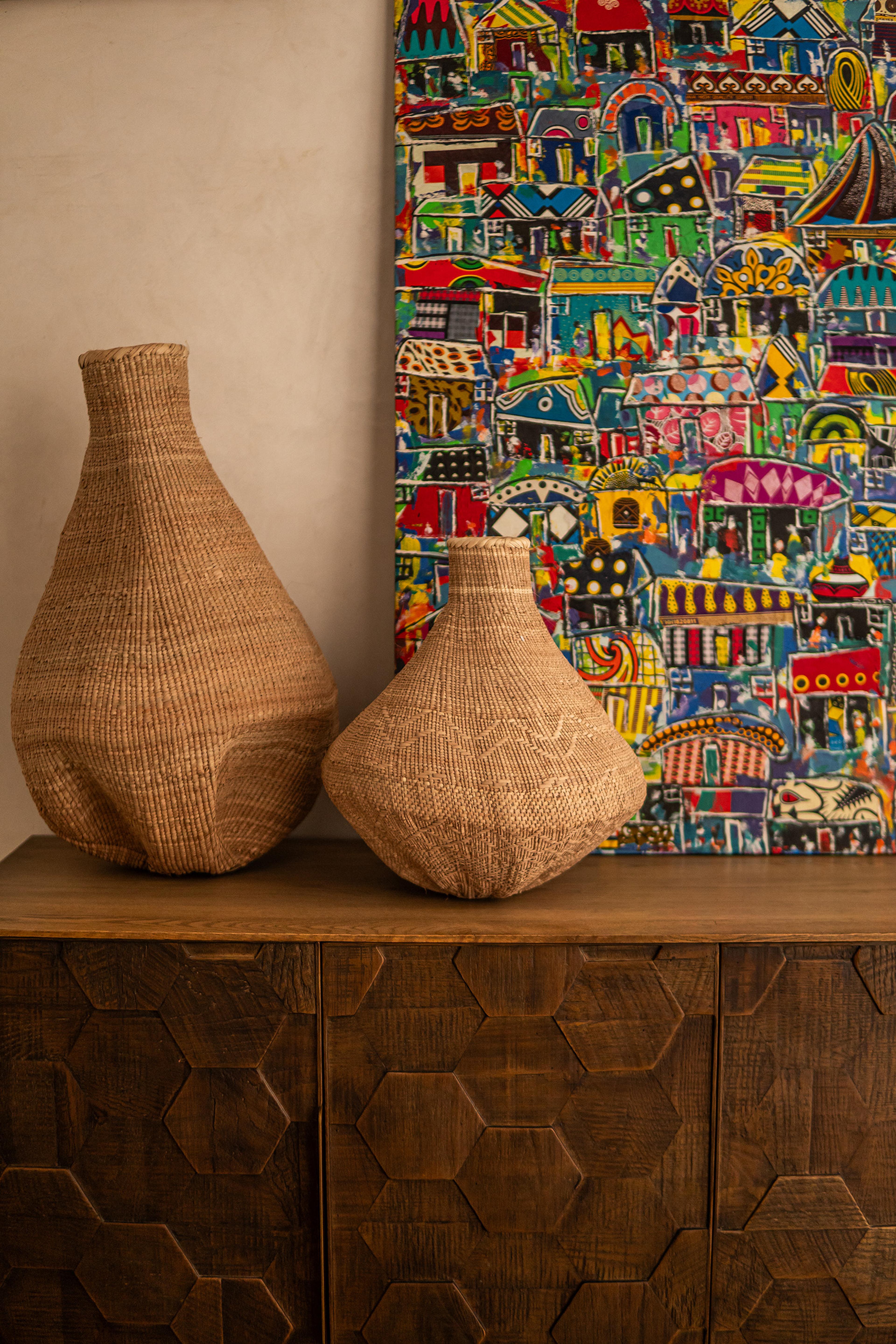Two large woven baskets stand on a carved wooden cabinet beside a colourful painting of houses.