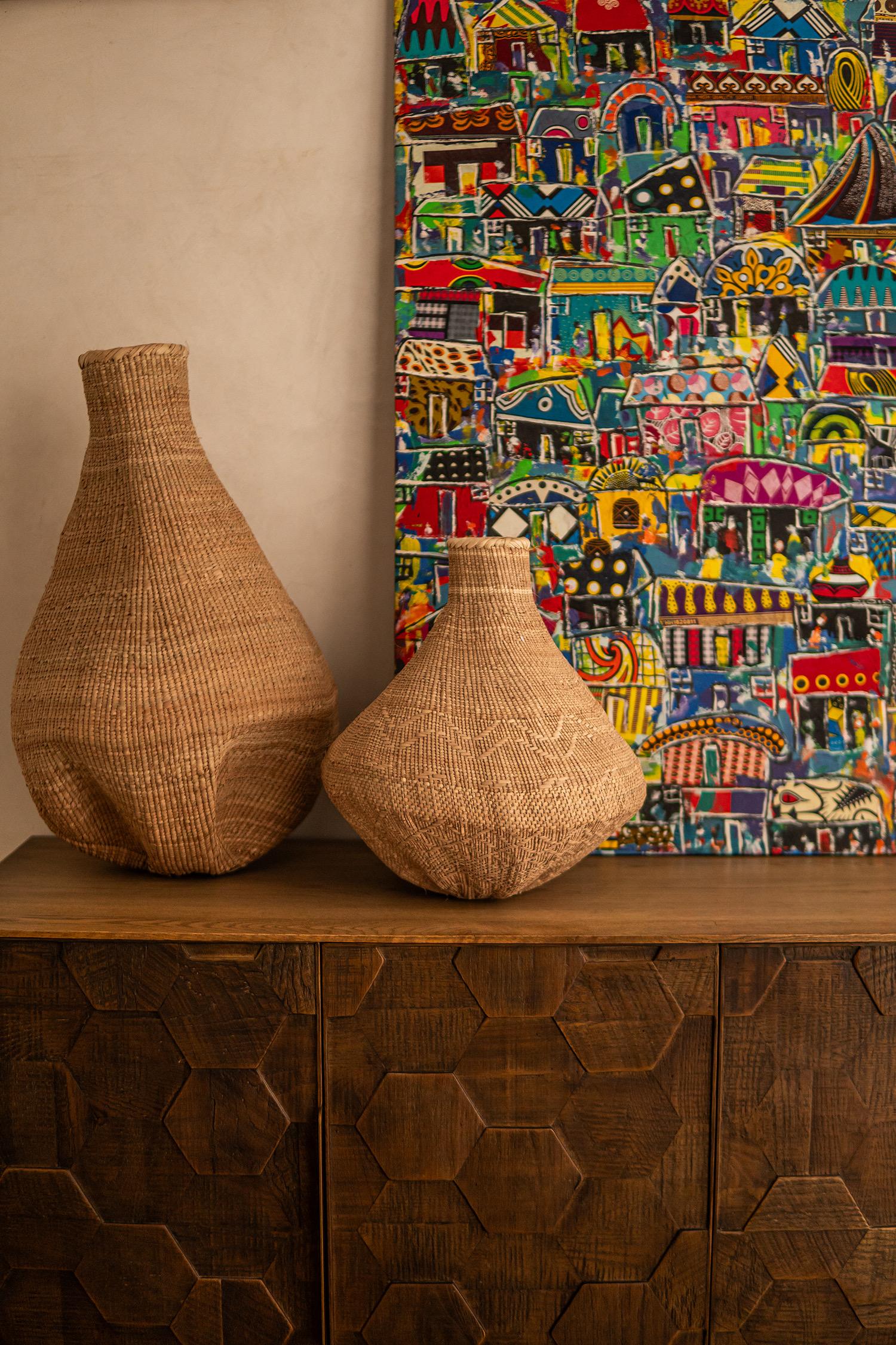 Two large woven baskets stand on a carved wooden cabinet beside a colourful painting of houses.