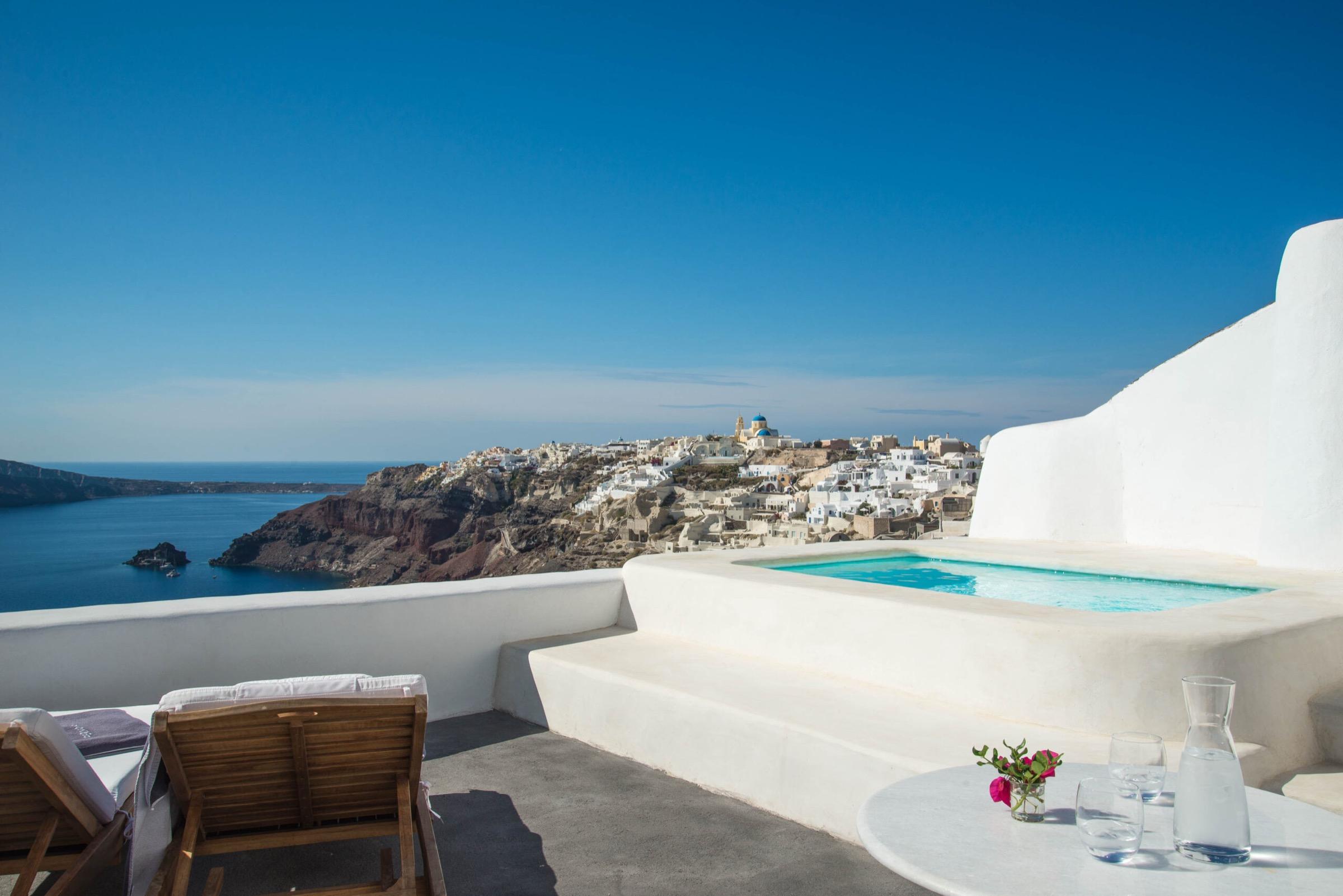 A plunge pool and wooden loungers on a whitewashed terrace, with a table set with a water jug and flowers, overlooking the sea and a cliffside village in the distance.