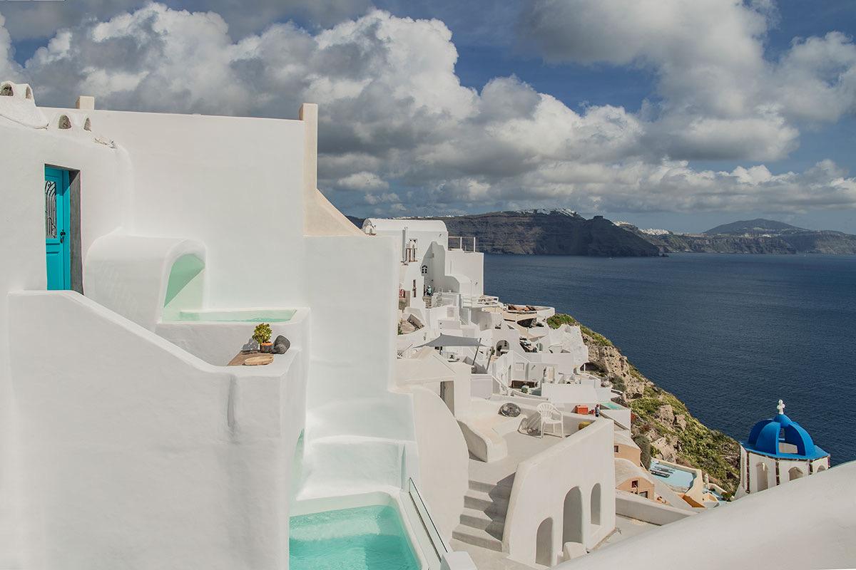 A white cubic building with a turquoise door sits on a cliff above the sea, with a plunge pool on a lower terrace and a blue-domed church beside it.