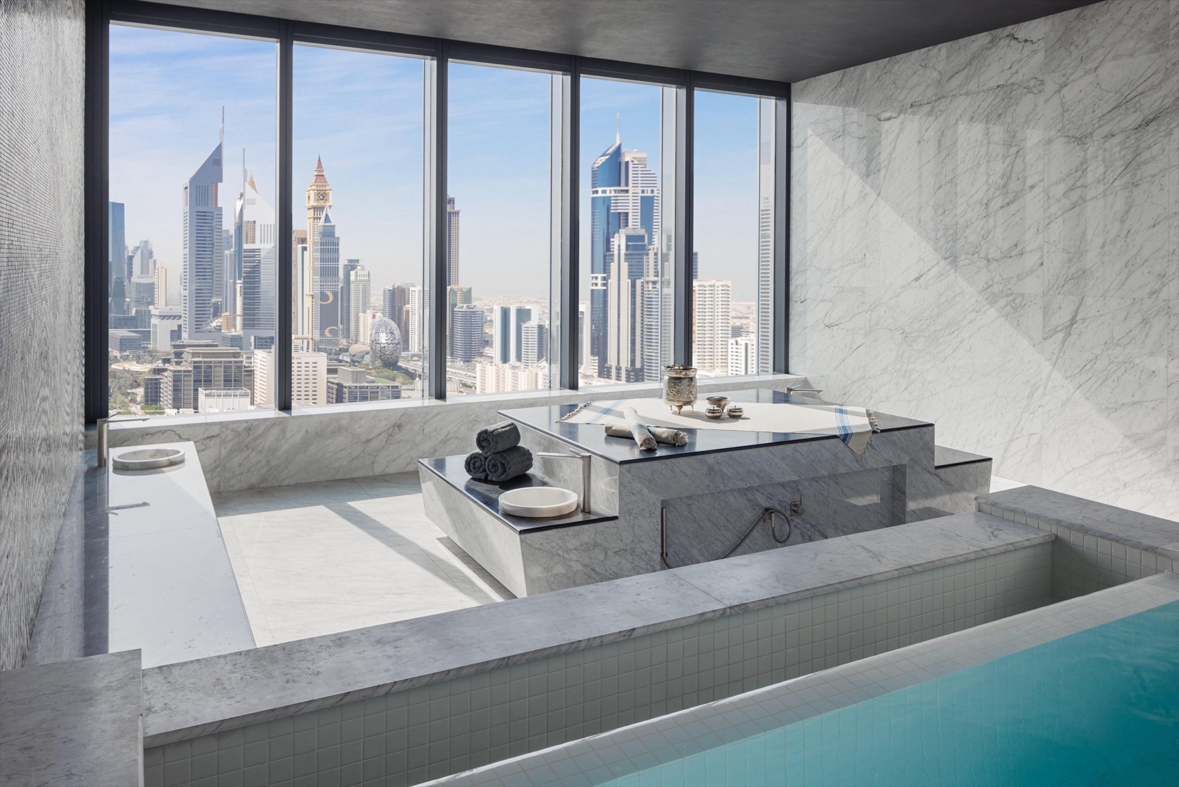 A marble bathing area with a tiled plunge pool in the foreground, a stone counter set with rolled towels and bath items, and a wall of windows looking over a dense skyline.