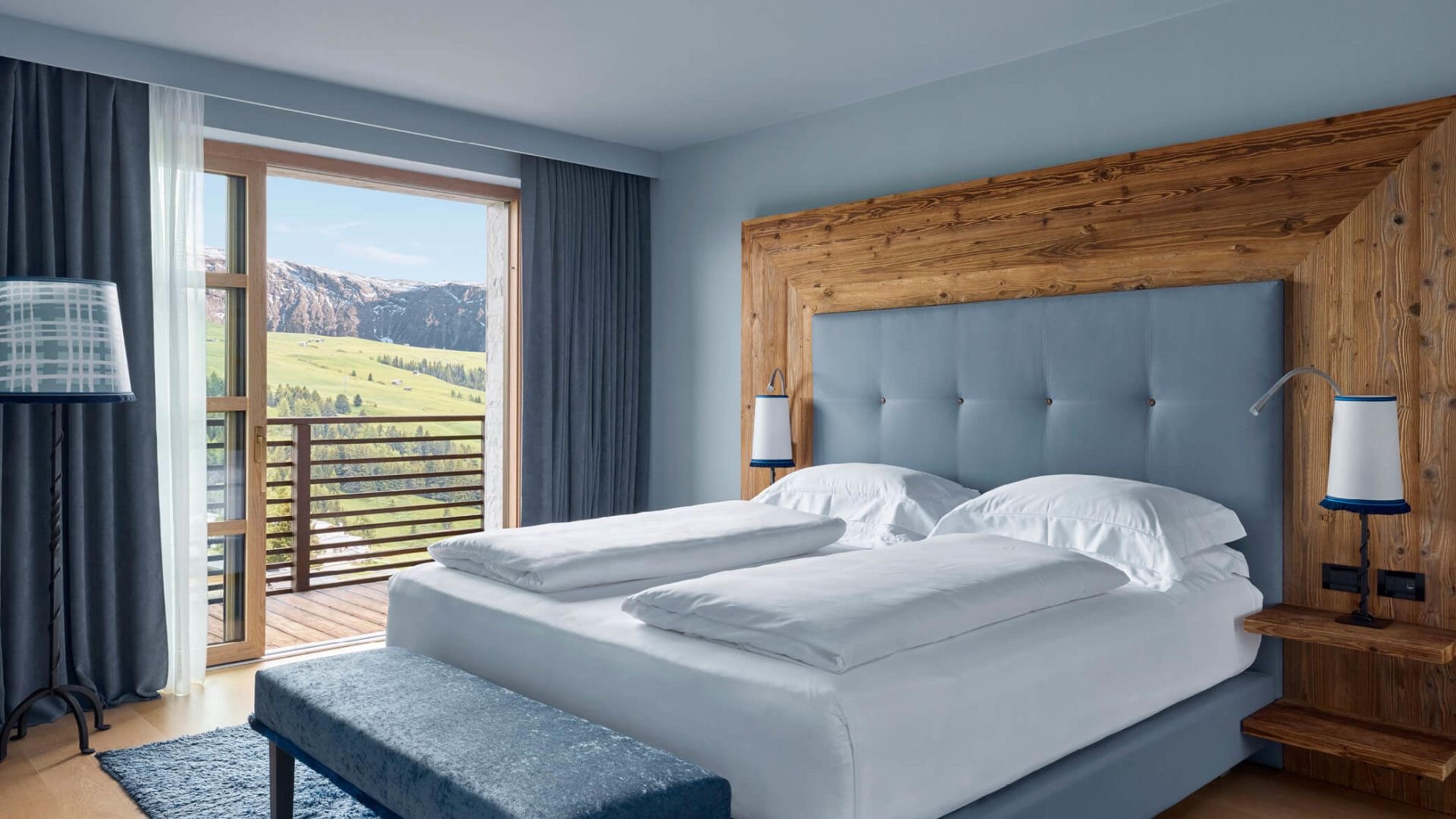 A bedroom with a tufted blue headboard built into a timber frame, its glass balcony doors framing a jagged grey peak across the valley.