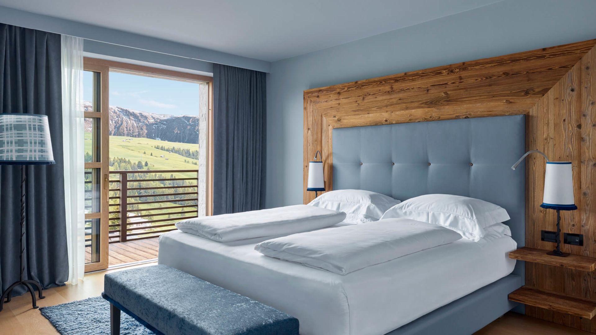 A bedroom with a tufted blue headboard built into a timber frame, its glass balcony doors framing a jagged grey peak across the valley.