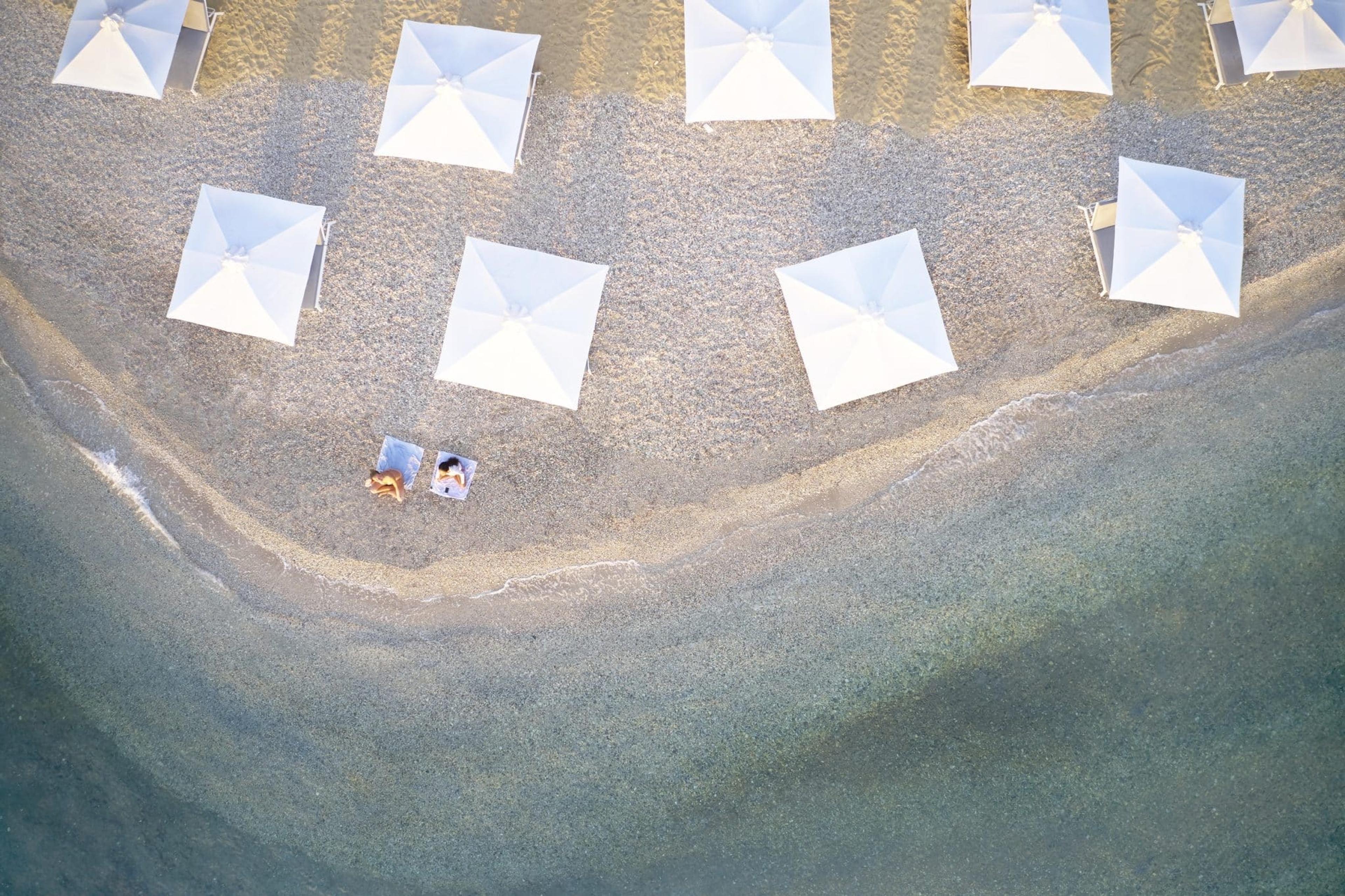 An aerial view of a shingle beach with rows of white umbrellas and two sunbathers lying on towels at the water's edge.