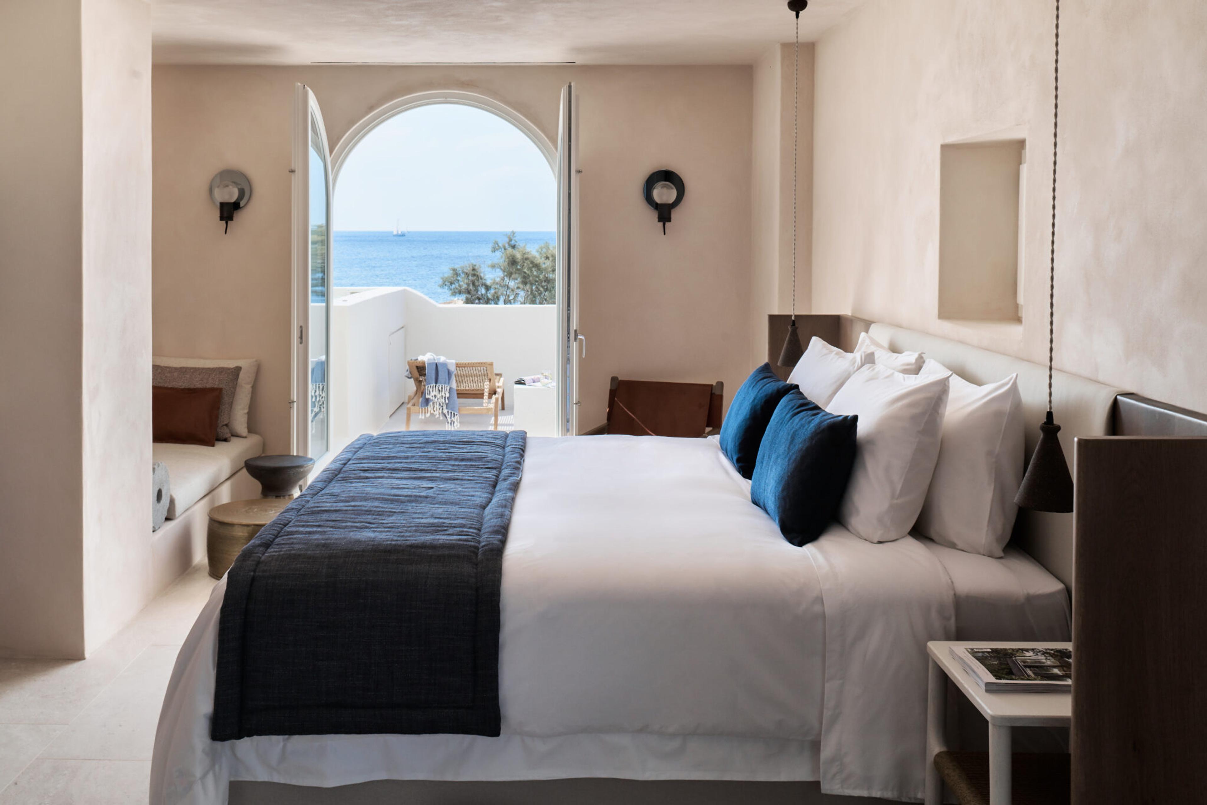 A bedroom with a dark blue throw and pillows on a white bed, facing an arched doorway open to a sea view terrace.
