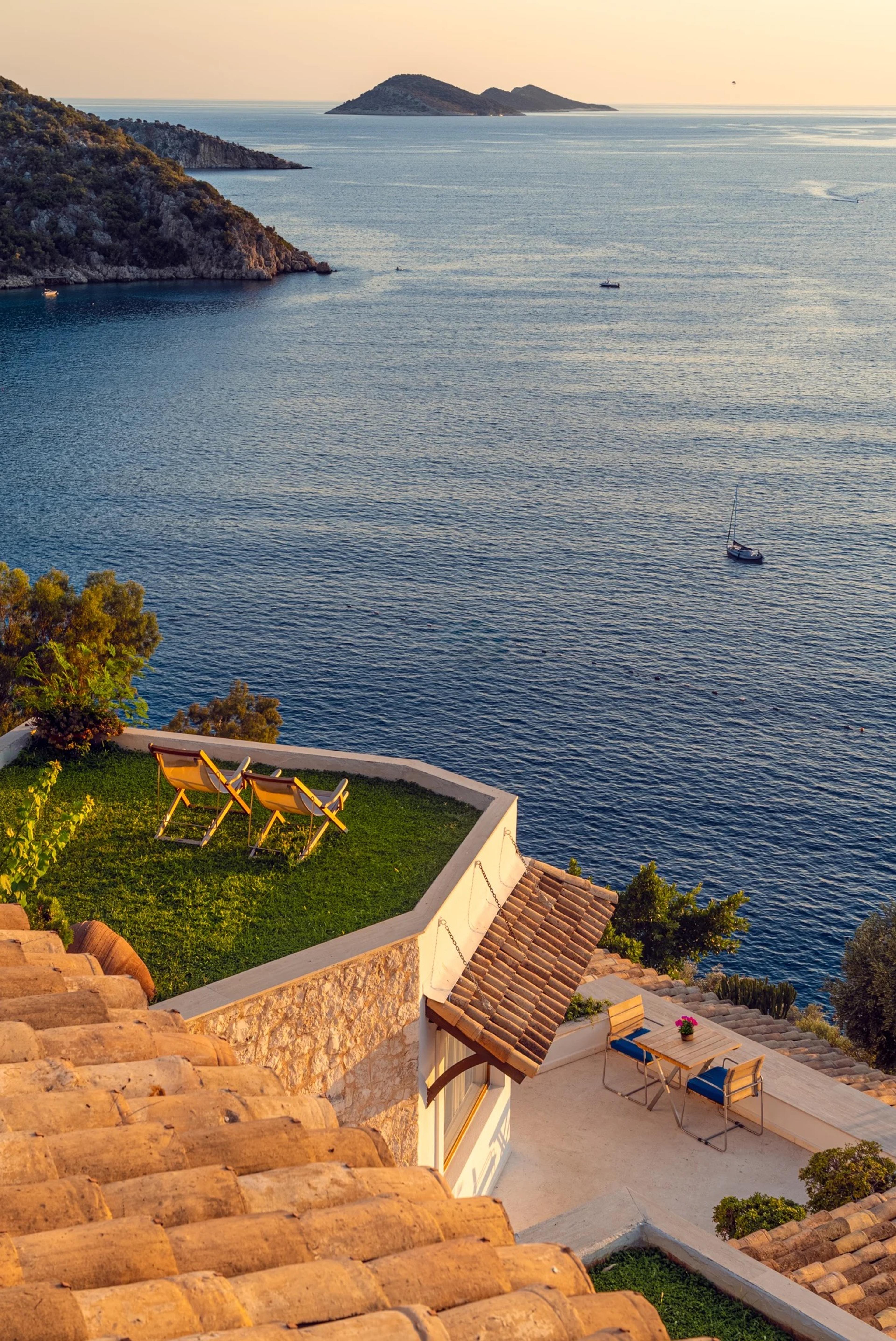 A tiled-roof building with a green lawn roof and two deck chairs, looking out over the sea to a rocky headland and an island in evening light.