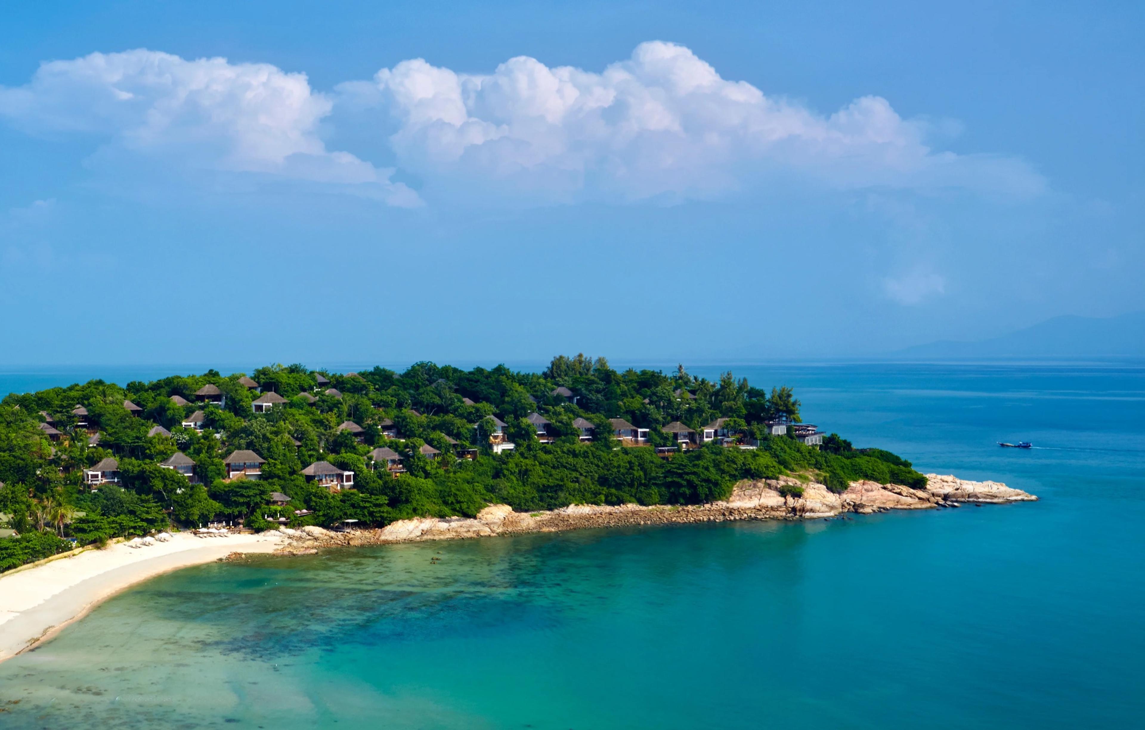 An aerial view of a green, tree-covered headland dotted with thatched roofs, bordered by white sand and turquoise water under a partly cloudy sky.