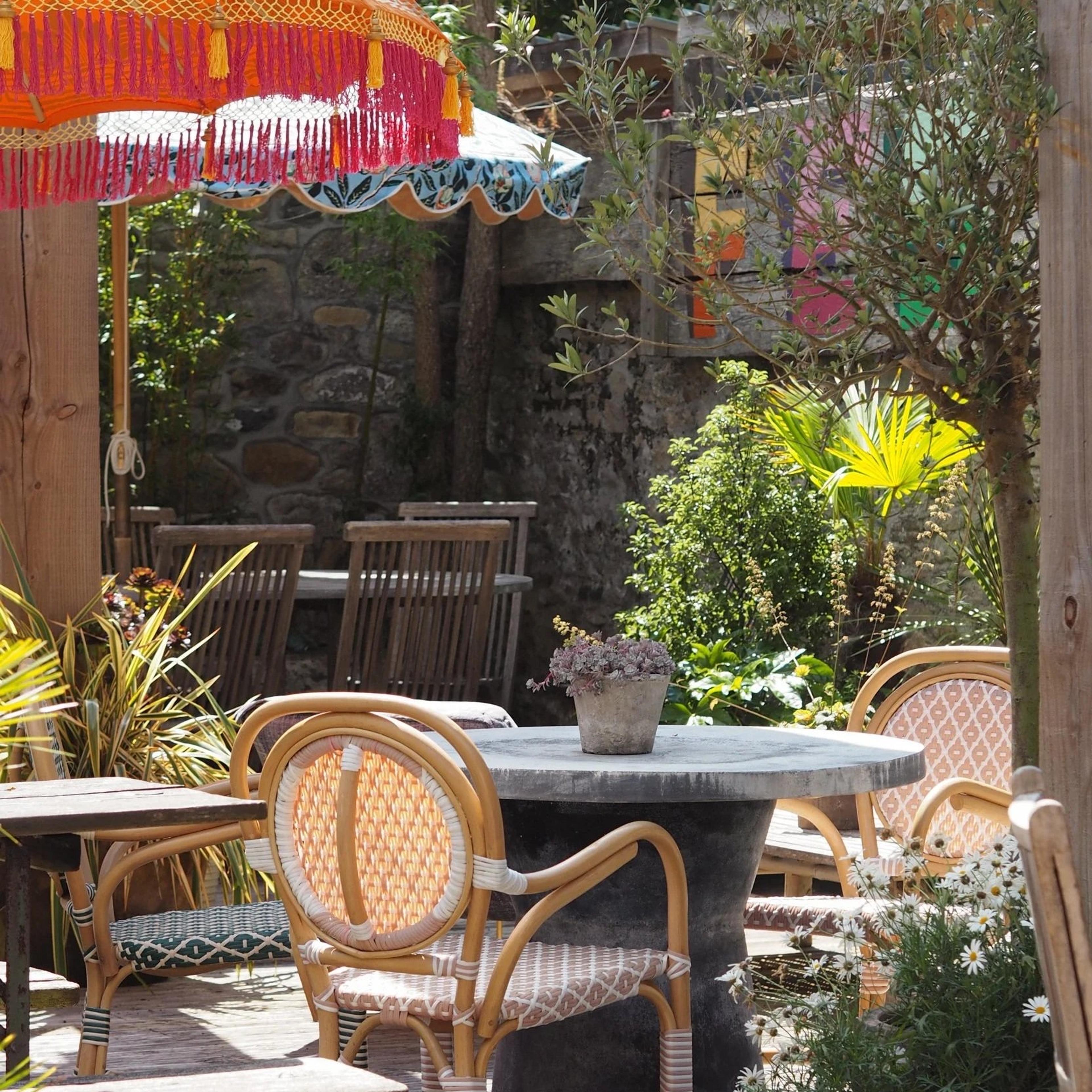 A sunlit terrace with rattan chairs around a stone table, a potted plant, daisies in bloom, and a red and orange patterned umbrella overhead.