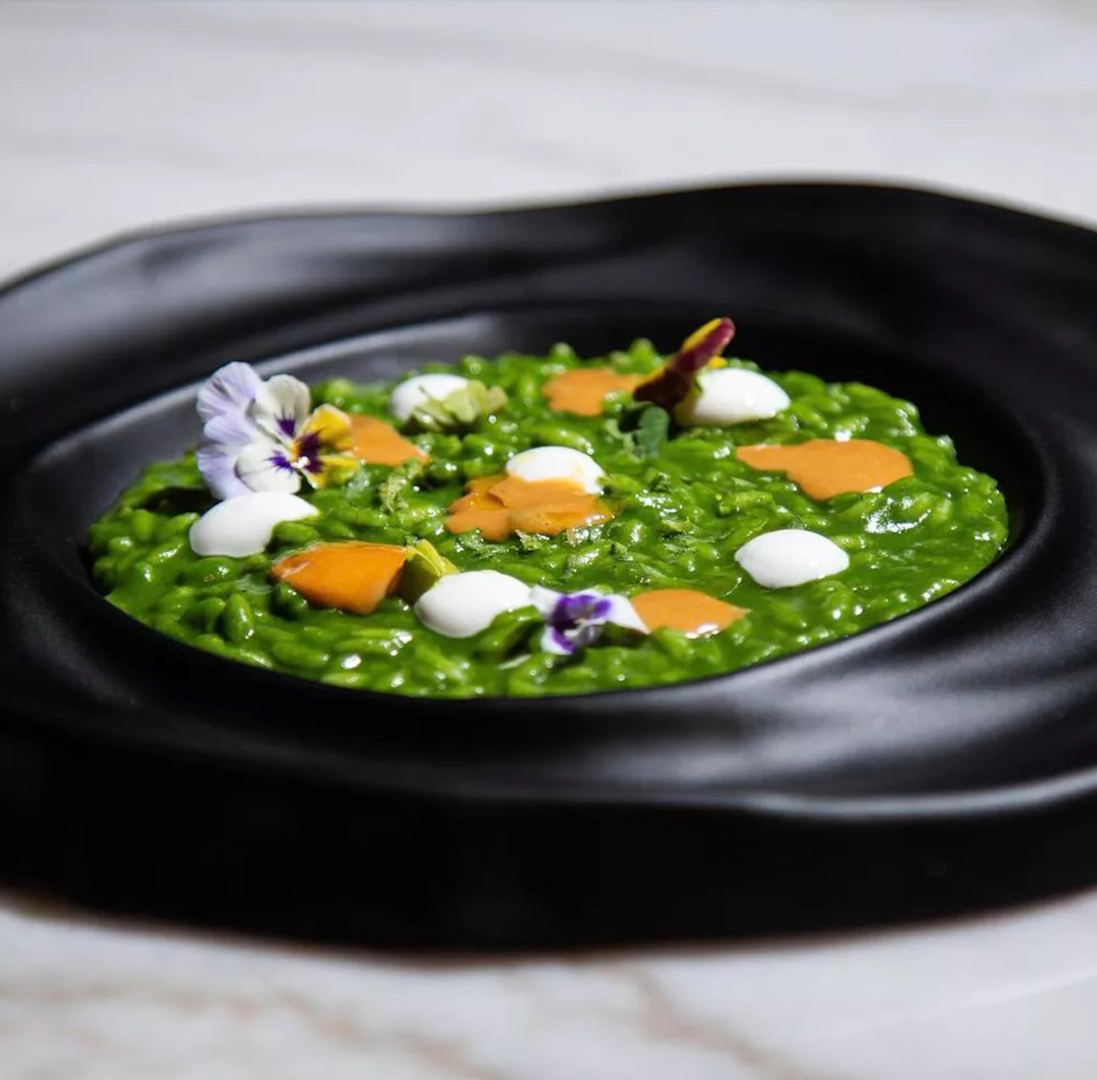 A plate of green risotto dotted with flowers and orange shavings on a black dish at MEC