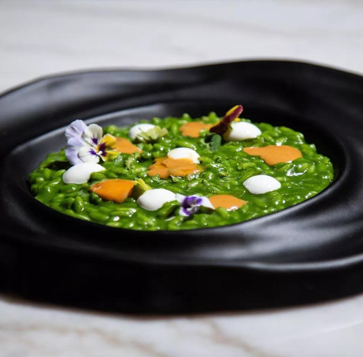 A plate of green risotto dotted with flowers and orange shavings on a black dish at MEC