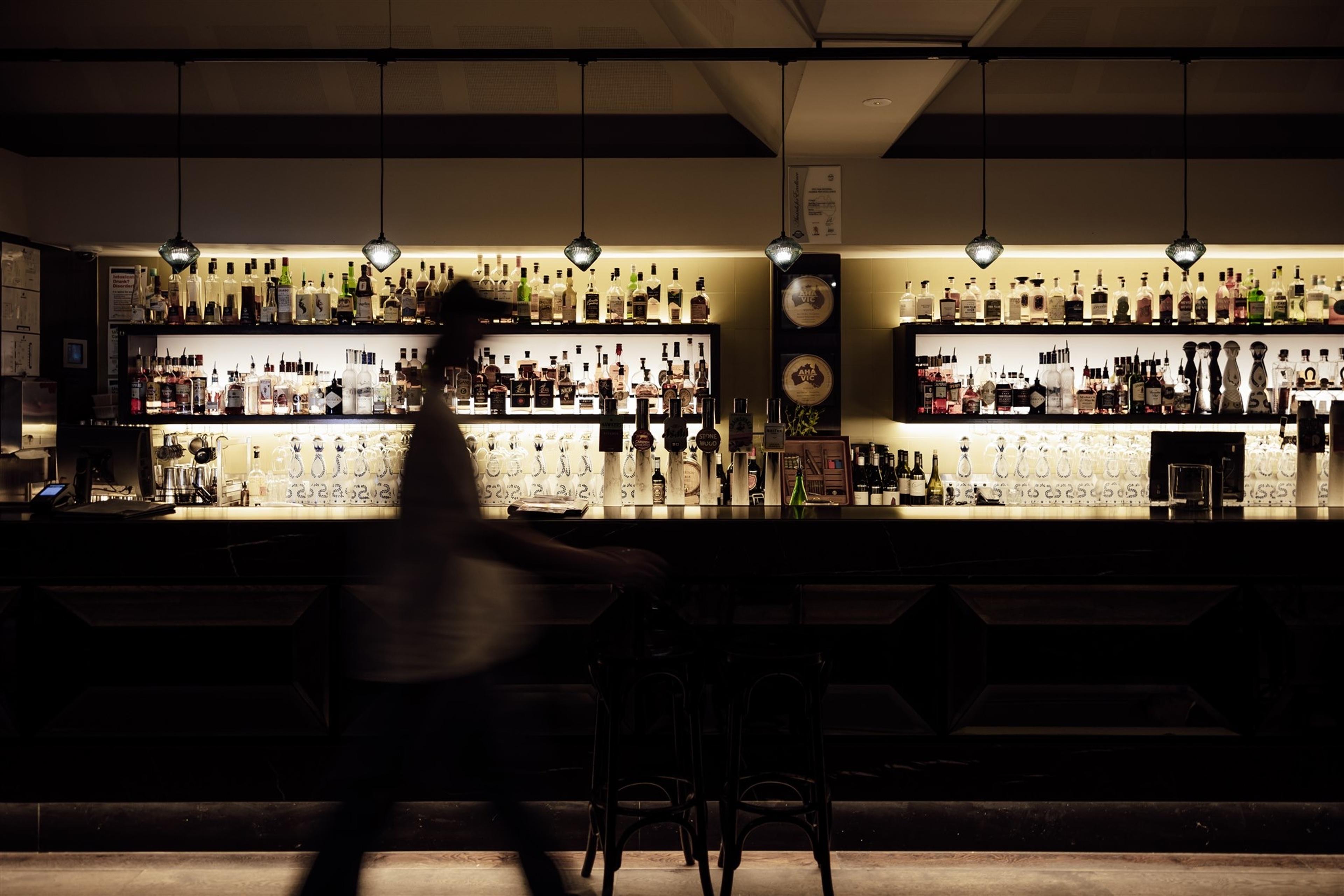 a dimly lit bar with rows of bottles on illuminated shelves and a blurred figure walking past the counter