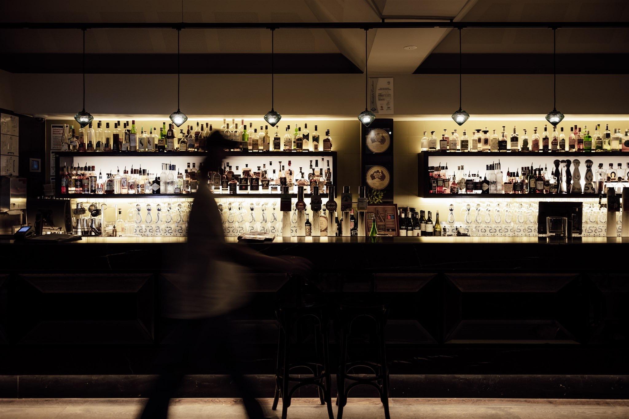 a dimly lit bar with rows of bottles on illuminated shelves and a blurred figure walking past the counter