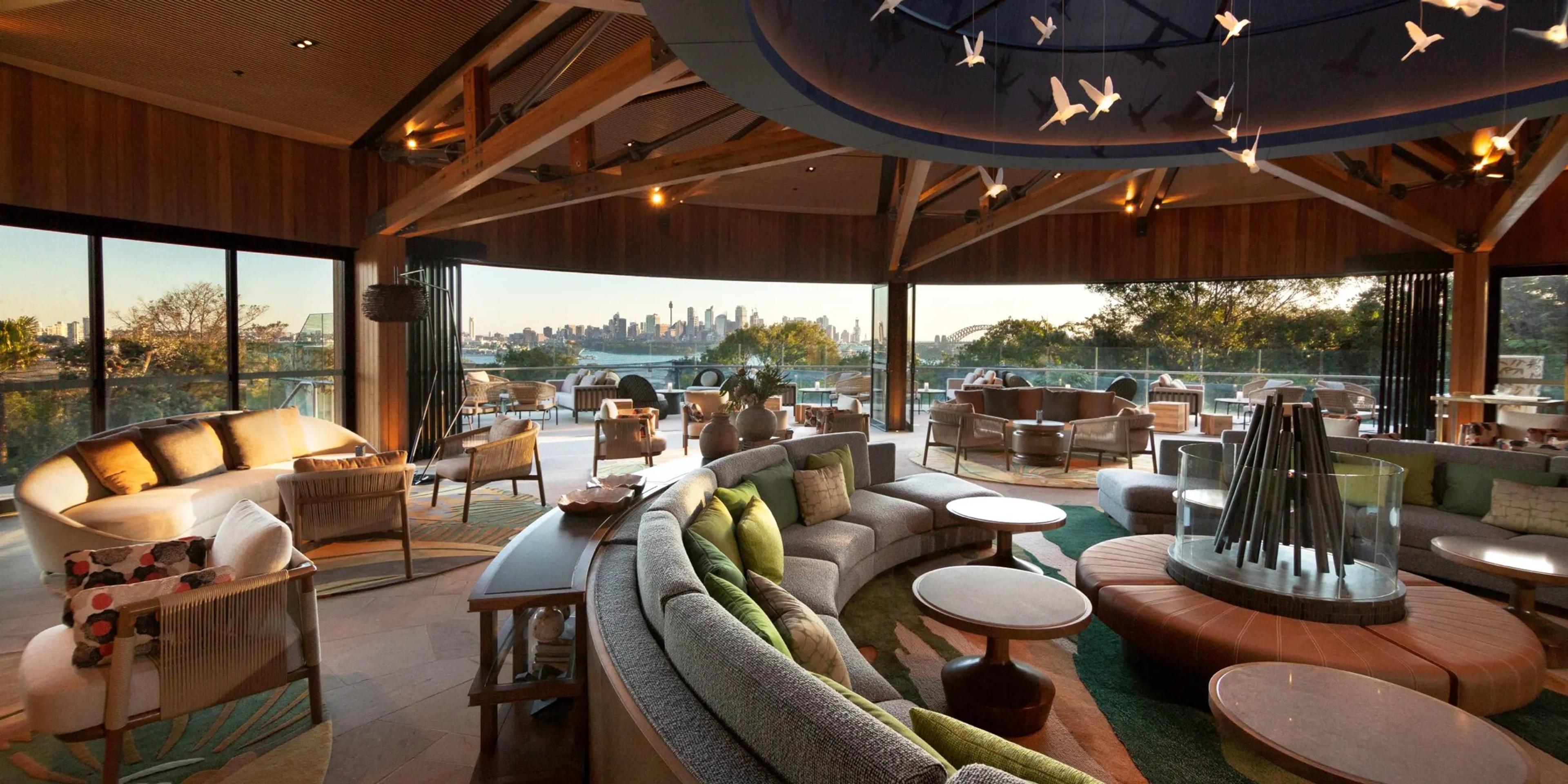 A curved cream sofa and low tables in a timber-beamed lounge, with a mobile of paper birds hanging from the ceiling and the Sydney skyline visible through floor-to-ceiling windows.