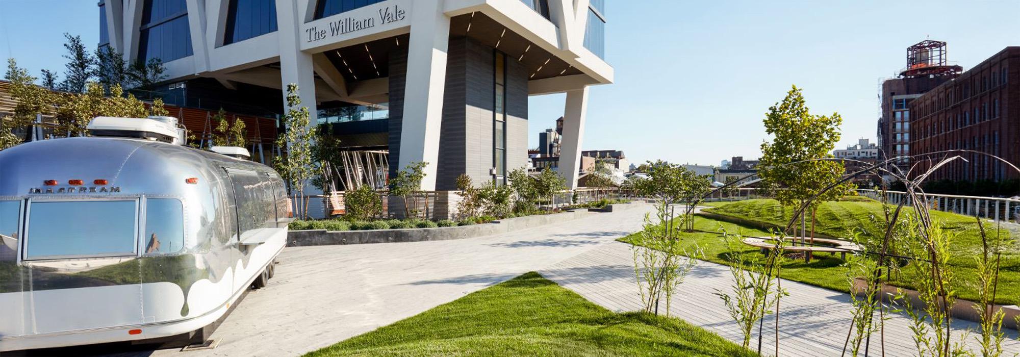 The William Vale's entrance at street level, with its name on the facade, a vintage silver trailer parked outside, and a landscaped garden path under a clear sky.