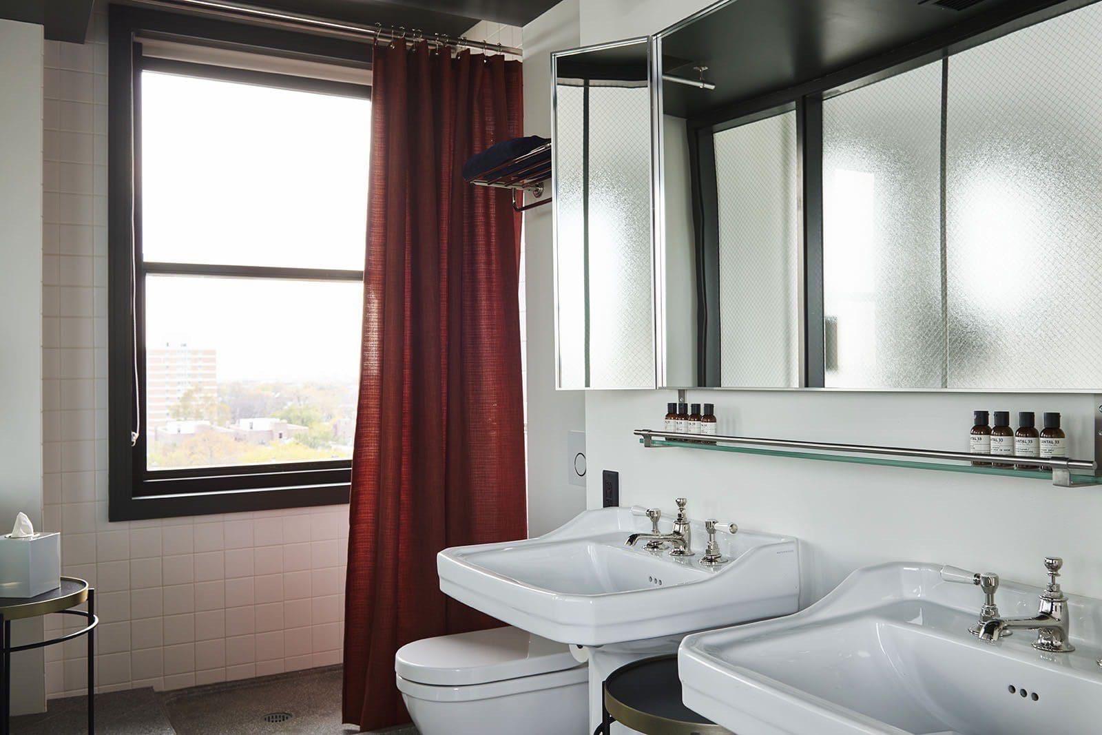 a bathroom with two white pedestal sinks under a black-framed mirror, a red curtain hanging by a window that shows the city beyond, and amenity bottles on a glass shelf